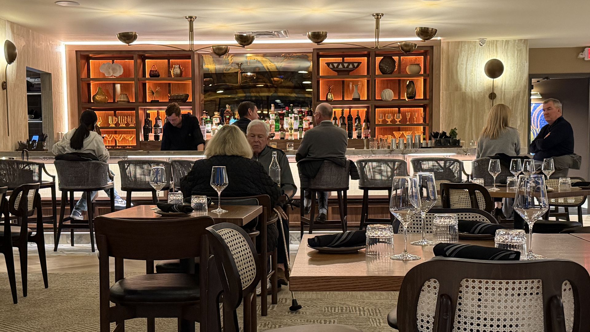 Interior of a cozy bar with patrons seated at tables and stools. Warm orange backlighting highlights shelves with bottles and decor behind the bar. Dark wood furniture and glassware adorn the tables.