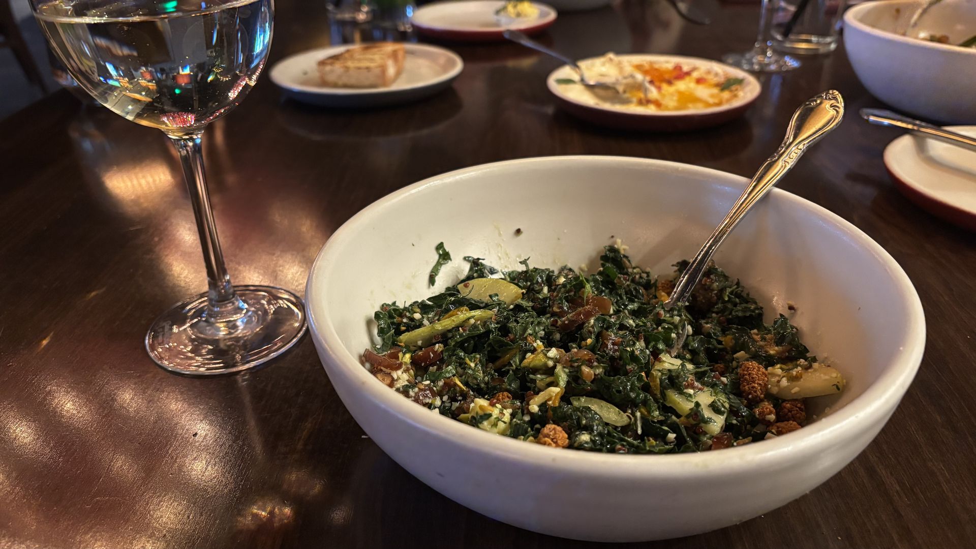 A white bowl of kale salad with nuts and feta on a dark wooden table, with a stemmed glass of white wine to the left and blurred plates in the background.