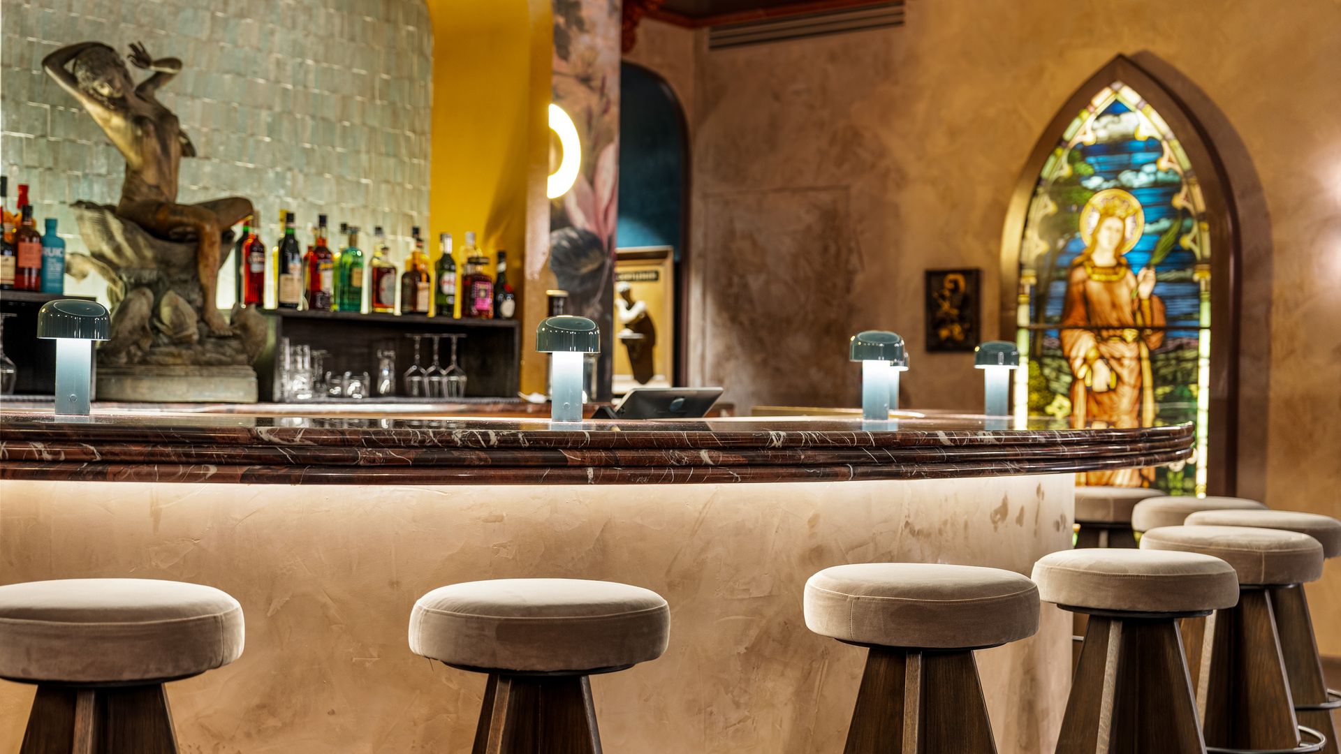 Elegant bar with brown marble countertop, cushioned wooden stools, small lamps, backlit bottles, a bronze sculpture, and a colorful stained glass window depicting a robed figure.