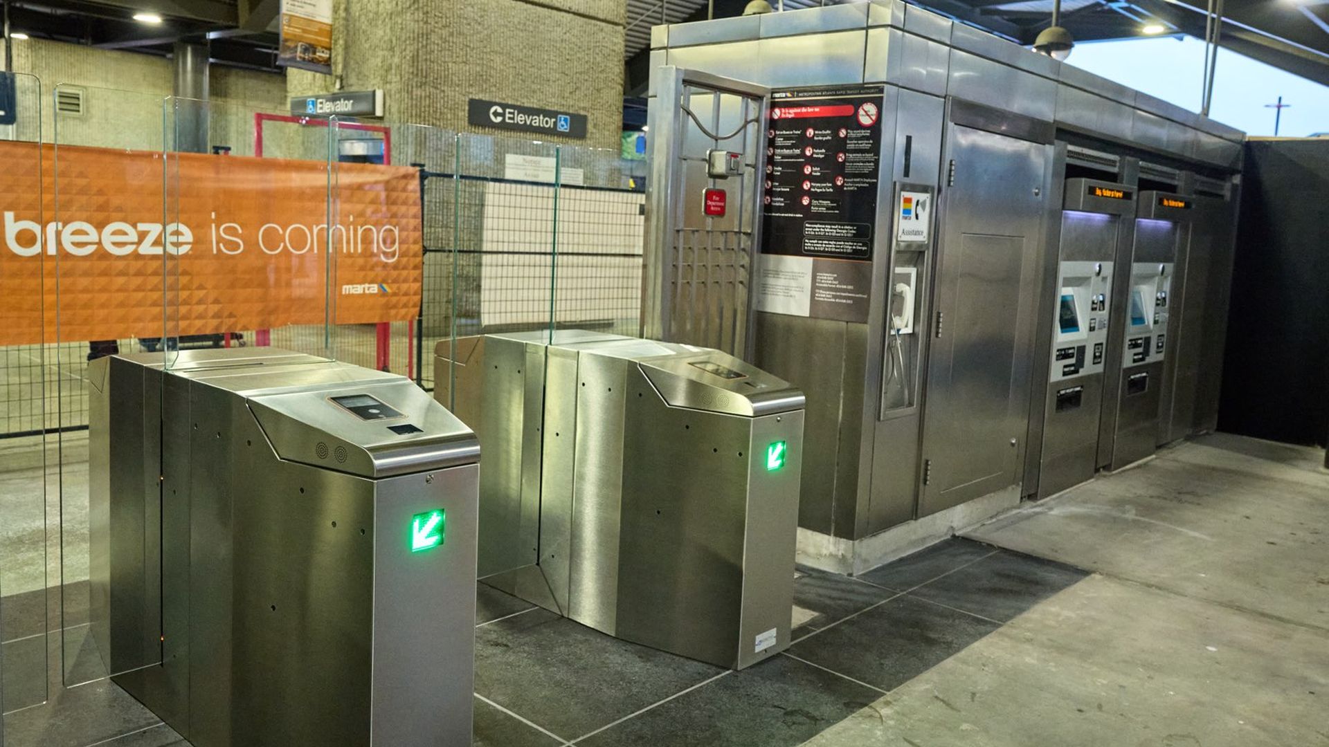 Modern metal subway turnstiles with green arrow lights, ticket machines on the right, and an orange sign saying "breeze is coming" in a transit station.