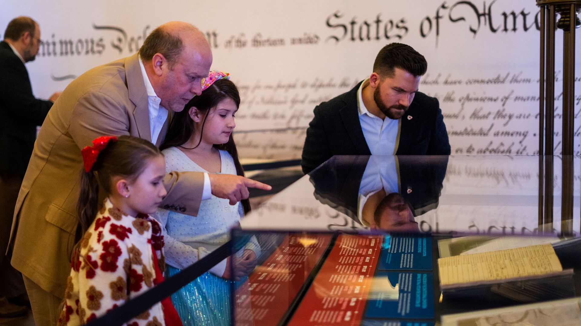 Family crowd at a museum display about the U.S. Constitution and founding documents. A bald man in a tan suit points to the exhibit while a girl in a blue dress and a bearded man look on at a glass case.