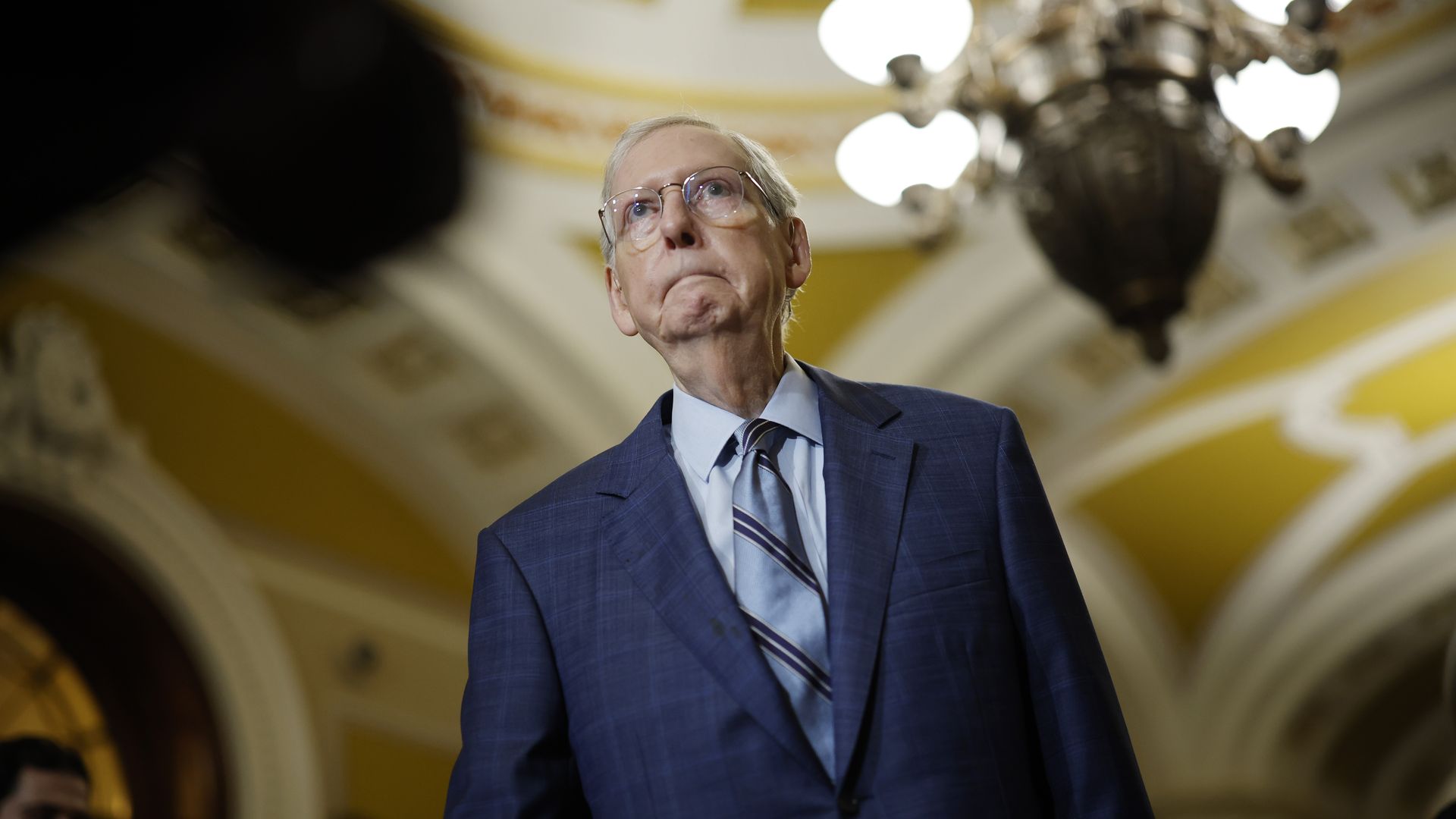 Mitch McConnell in a blue suit and striped tie. Photo is taken from below and a light fixture is in the background. The walls are yellow.