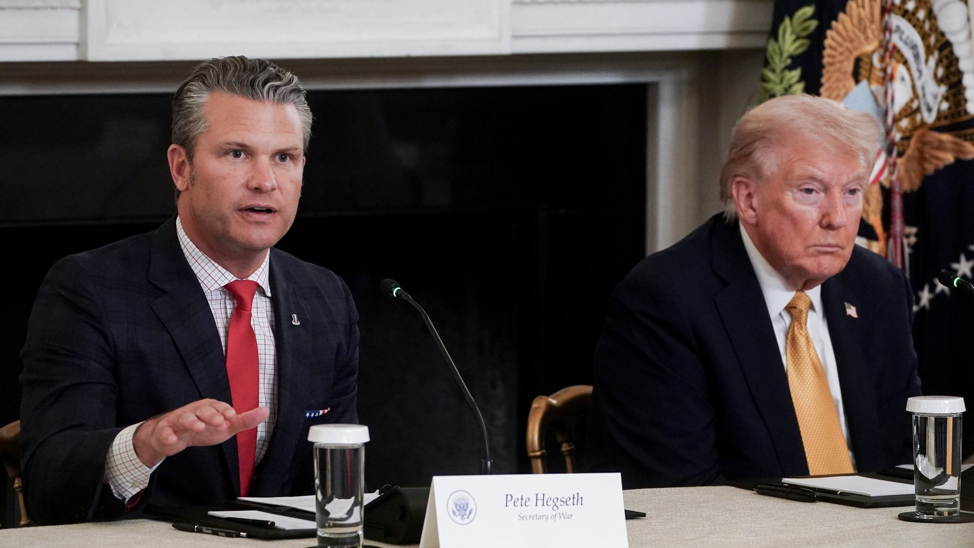 Pete Hegseth and Donald Trump, wearing dark suits and sitting at a table with glasses of water in front of a white fireplace.