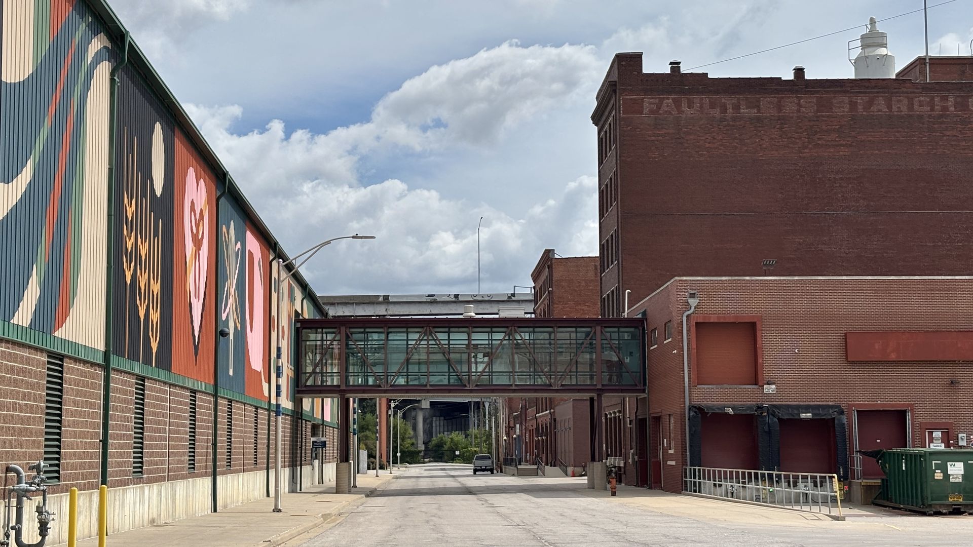 Urban alley flanked by brick buildings. The left wall features a bright mural with large flowers and bold colors; a glass footbridge links to the right building beneath a cloudy sky.