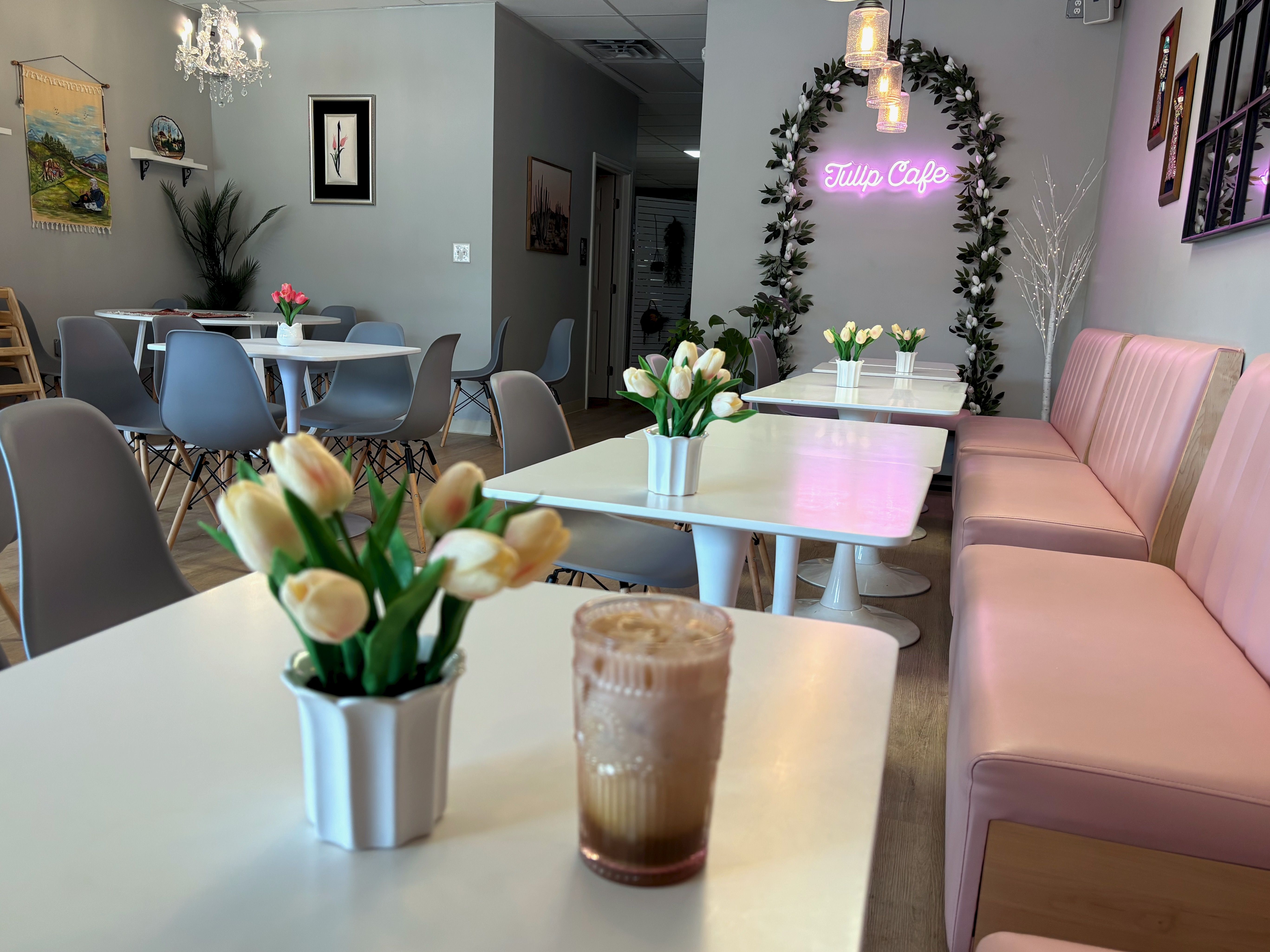 Pastel cafe interior with pink booths, white tables, and gray chairs. Tulip centerpieces on each table; a neon pink "Tulip Cafe" sign sits in a leafy floral arch by a chandelier.