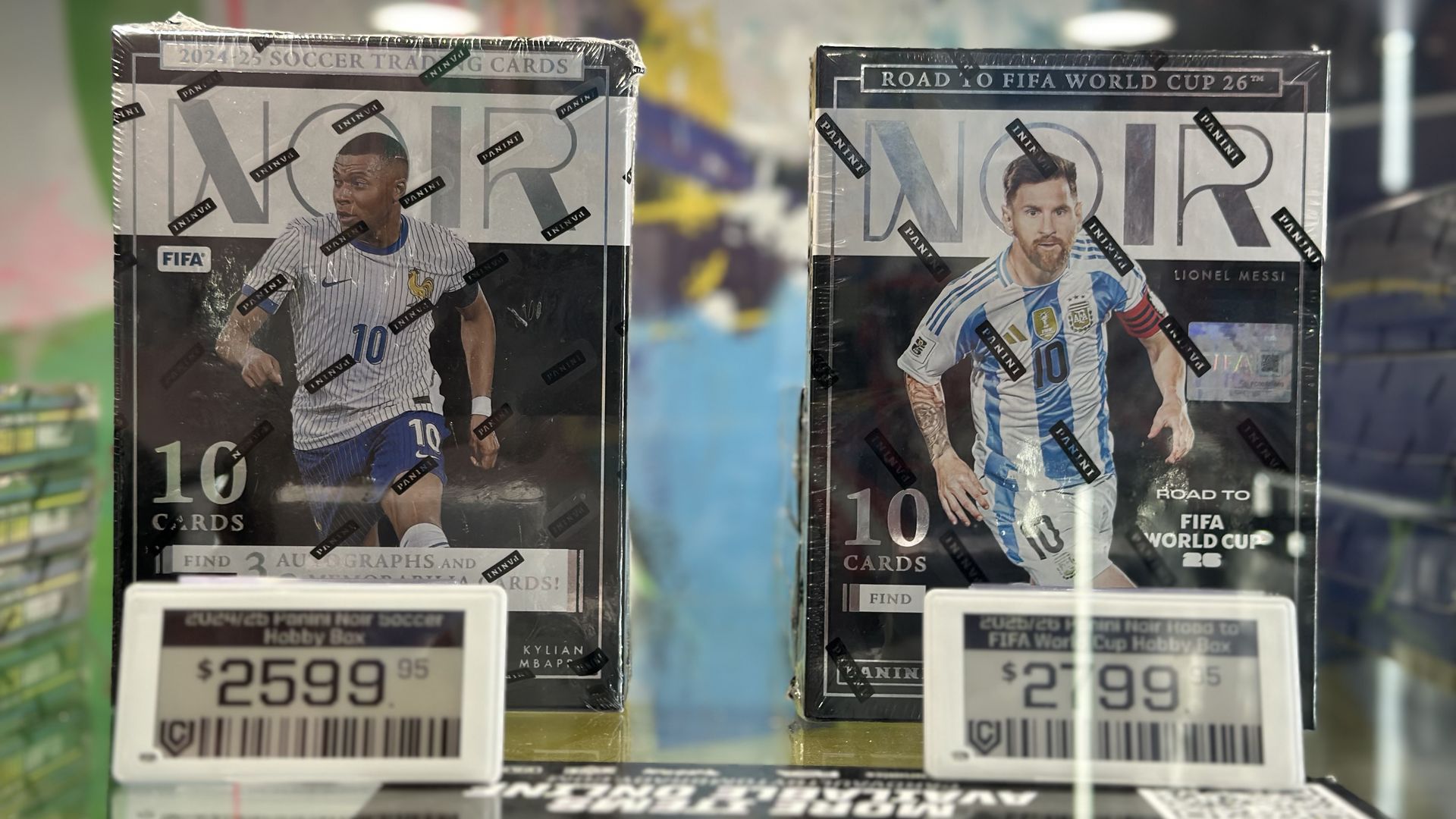 Two Panini FIFA World Cup trading card boxes on a store shelf, with France's Kylian Mbappé on the box cover to the left for $2,599, and Argentina's Lionel Messi on the box cover to the right for $2,799.