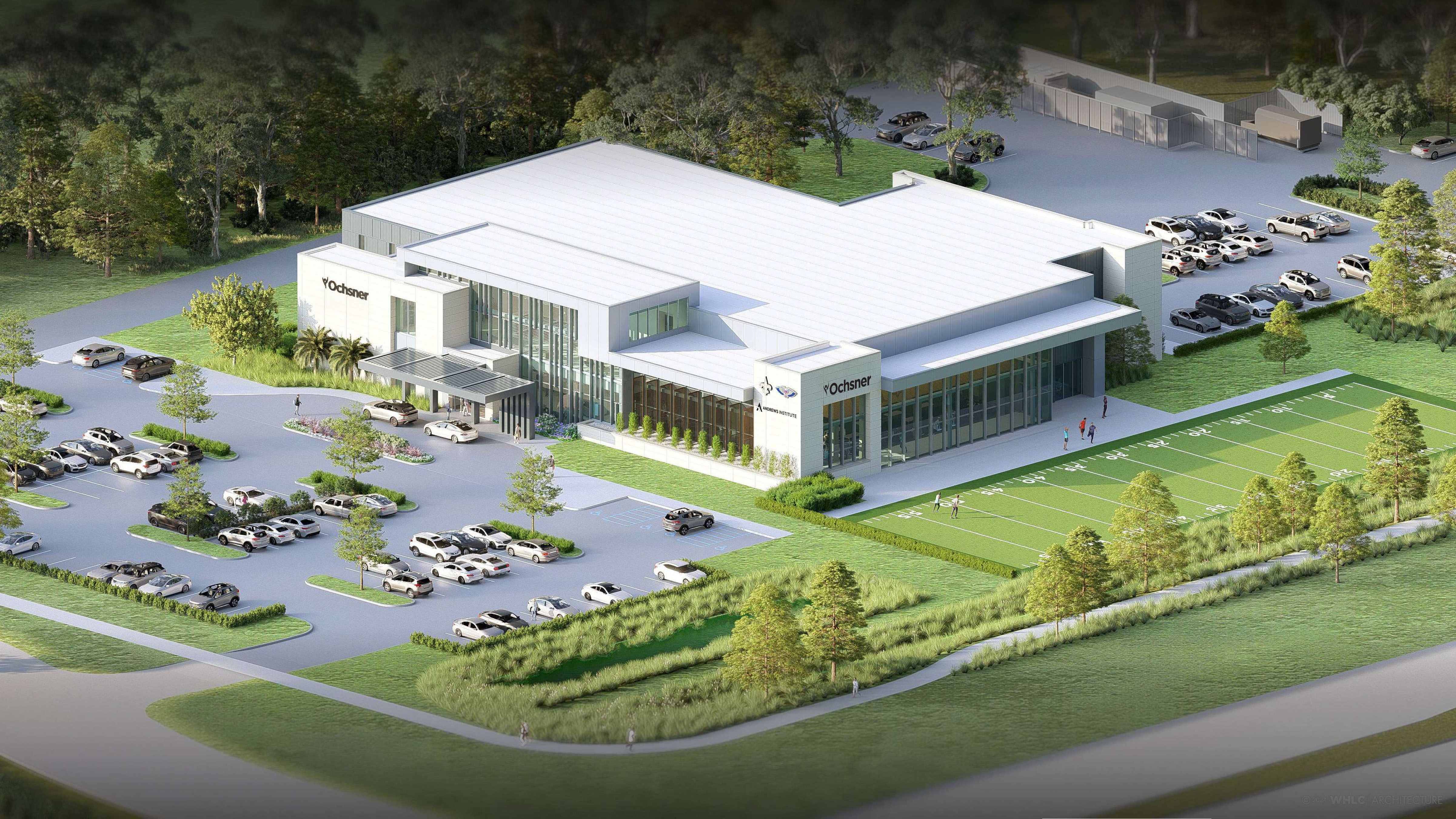 Aerial view of a modern white building labeled "Ochsner" with large glass windows, surrounded by green trees, a parking lot with cars, and an adjacent sports field marked with yard lines.