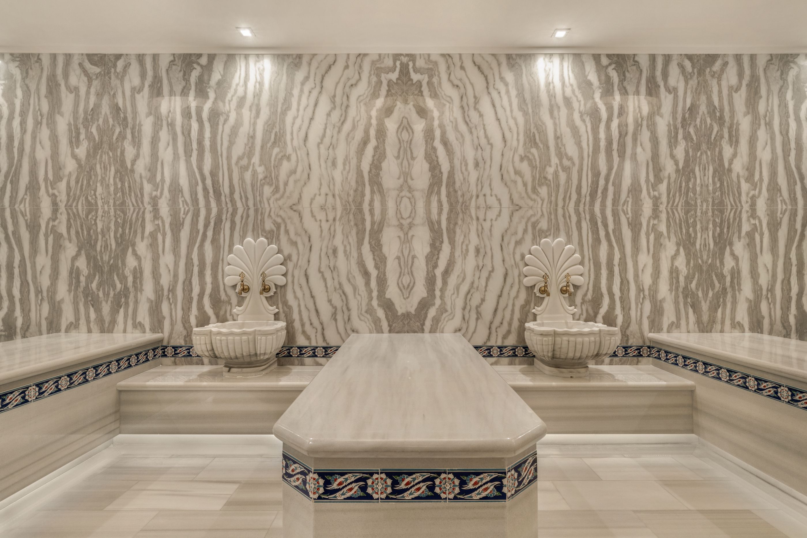 Luxurious spa room with beige-gray marble walls in bold vertical patterns, two white shell-shaped faucets on ornate pedestals, and a central marble table; blue tile border along the base.