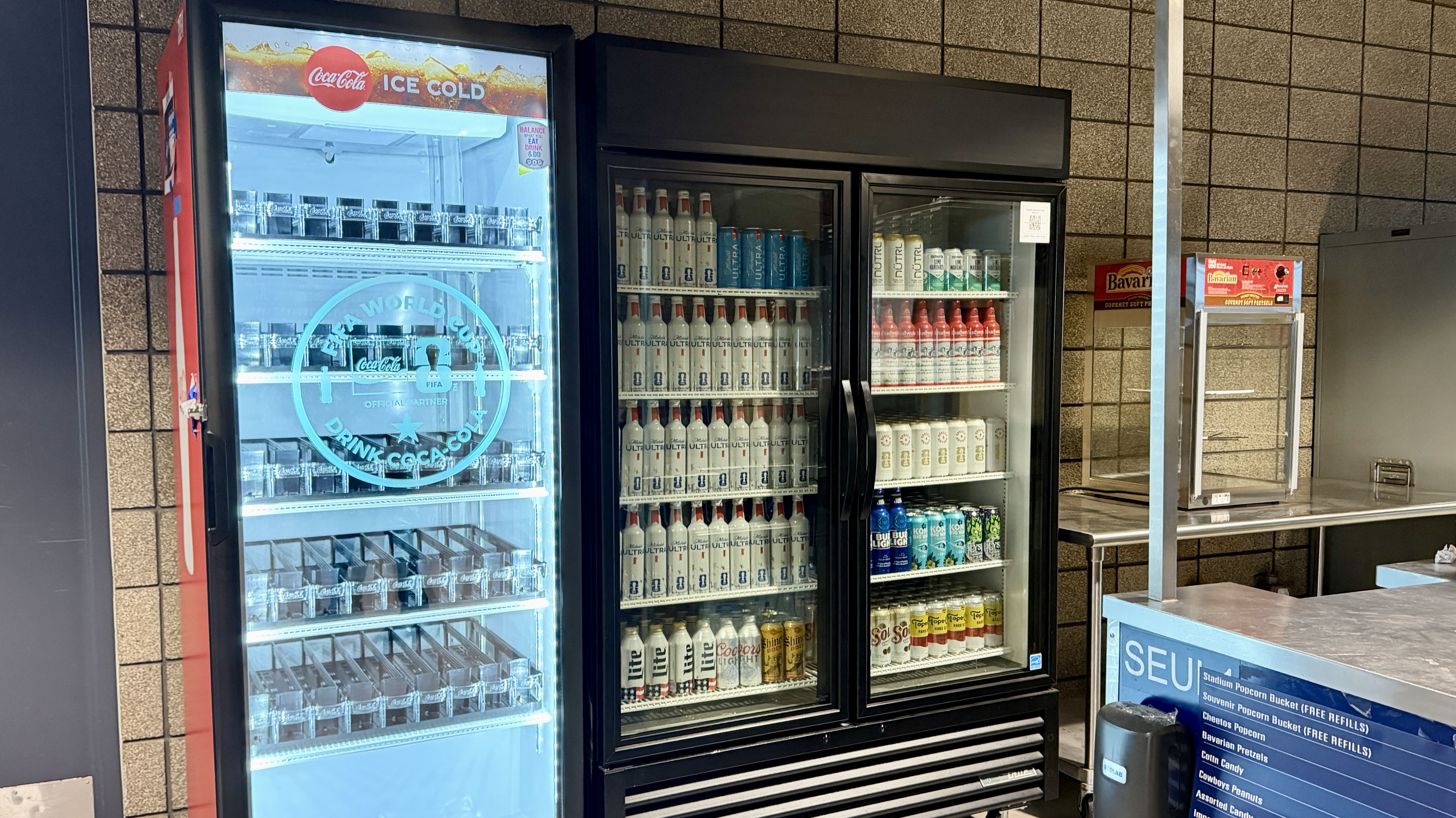 Three glass-front beverage fridges along a tiled wall: left blue-lit Coca-Cola Ice Cold cooler, middle ULTRA cans, right assorted bottles; Bavaria snack stand and stainless counter to the far right.