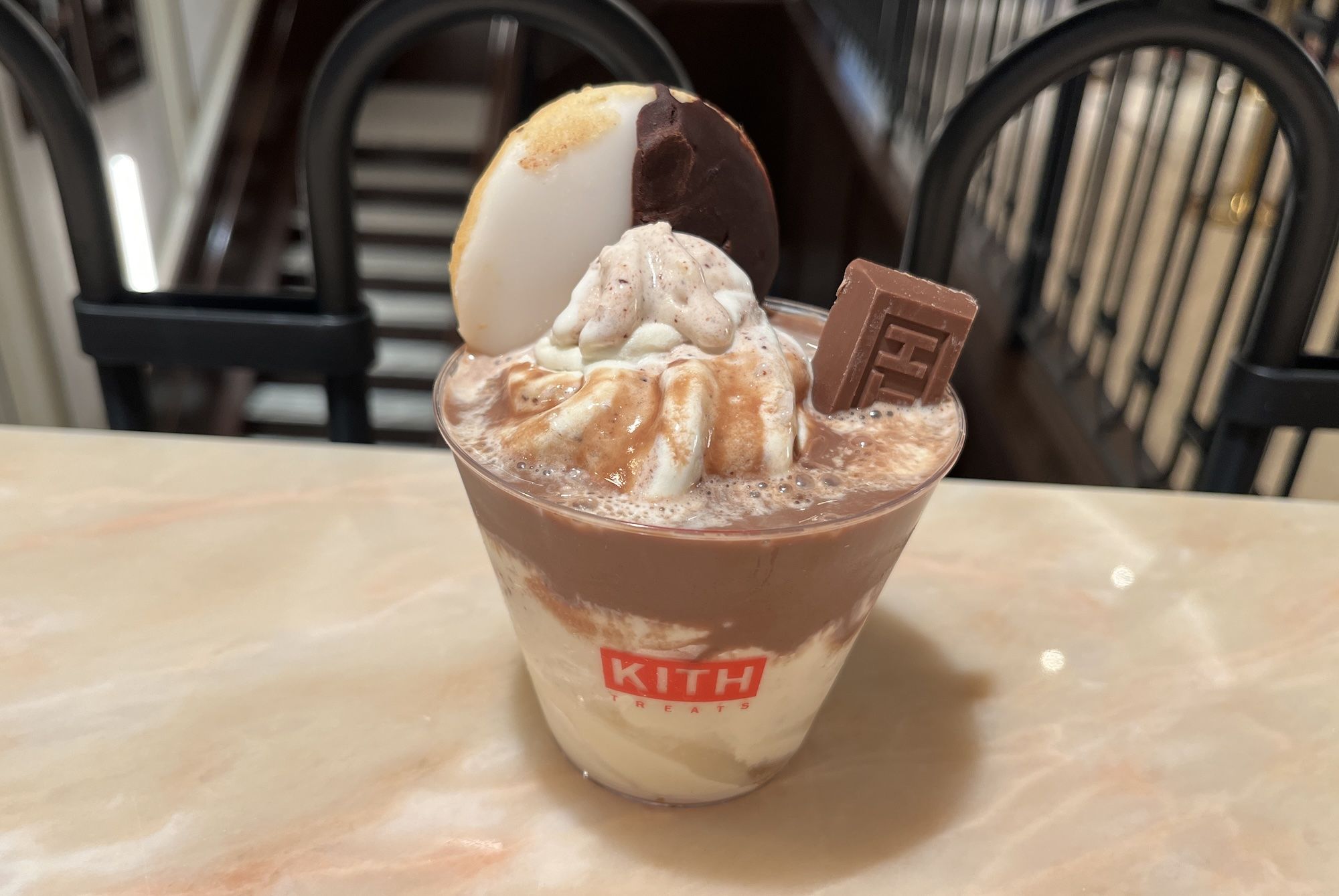Chocolate milkshake in a clear cup topped with whipped cream, caramel swirl, a round white cookie, and a chocolate square; a red KITH logo is visible.