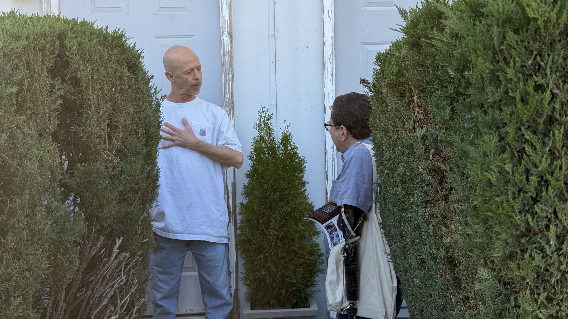 Two men stand by a weathered pale blue door between dense shrubs. The bald man in a light gray shirt places a hand on his chest; the other man with glasses holds a bag and a magazine.