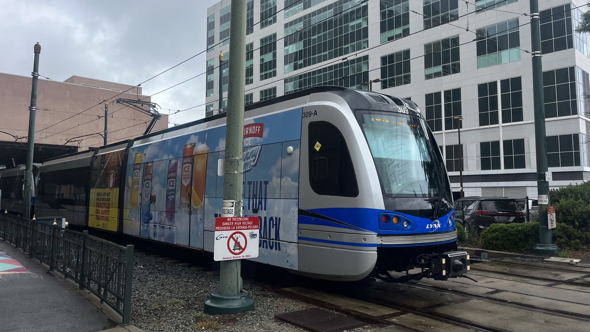 Blue and white Lynx light rail train on tracks in urban area with tall glass building in background, under cloudy sky. Train features advertisement with beer.