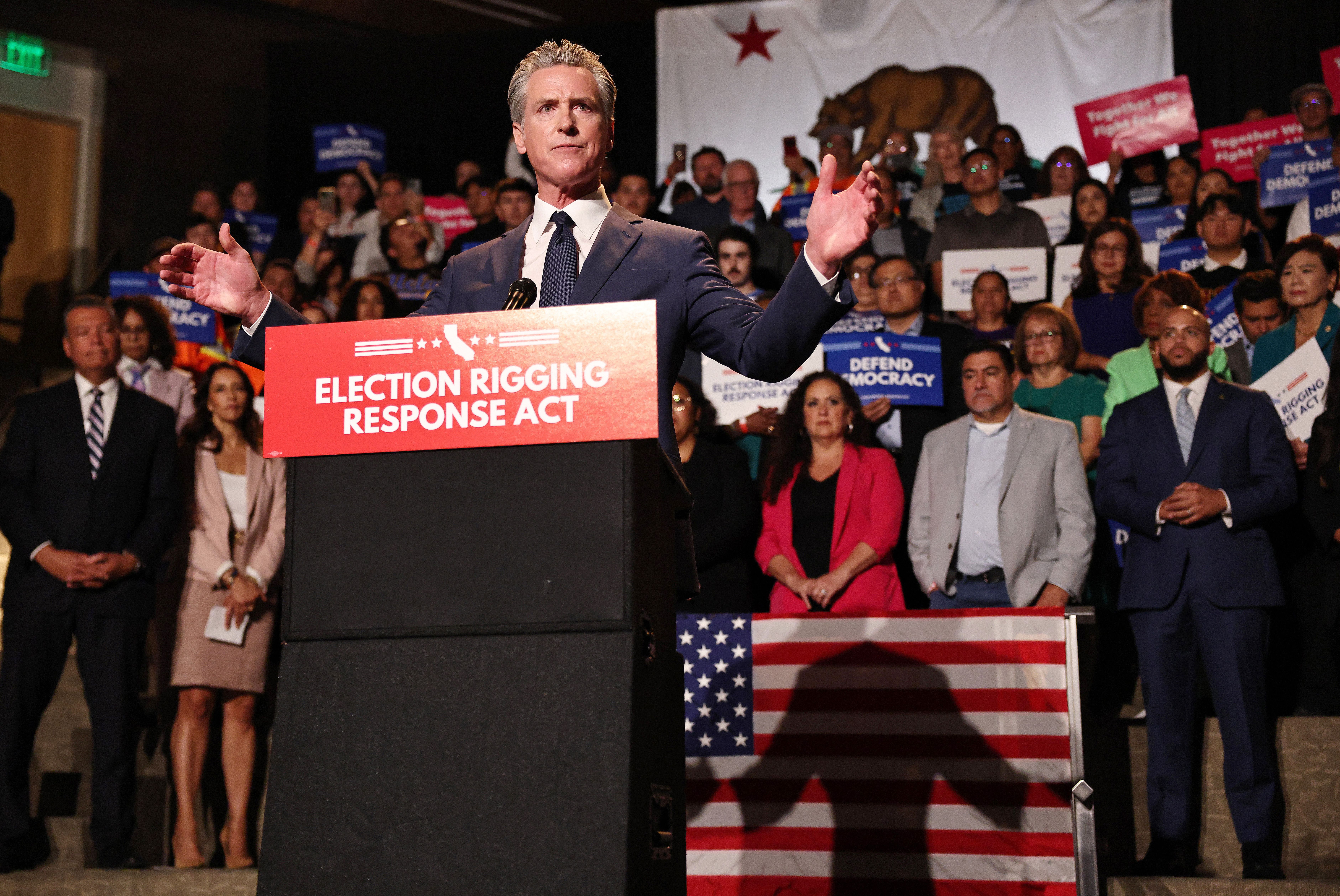 A man in a suit speaks at a podium with a red sign reading "ELECTION RIGGING RESPONSE ACT". Behind him, a crowd holds signs like "DEFEND DEMOCRACY" under a California flag.