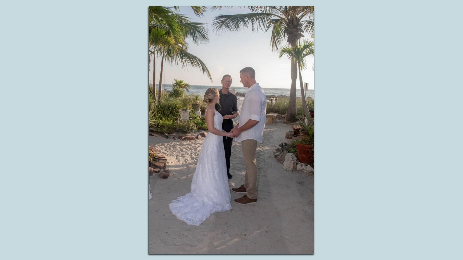 A woman in a wedding dress holding hands with a man in a white button-down and khakis. An officiant in black stands behind them. In the background is a lush garden, sandy beach and water.