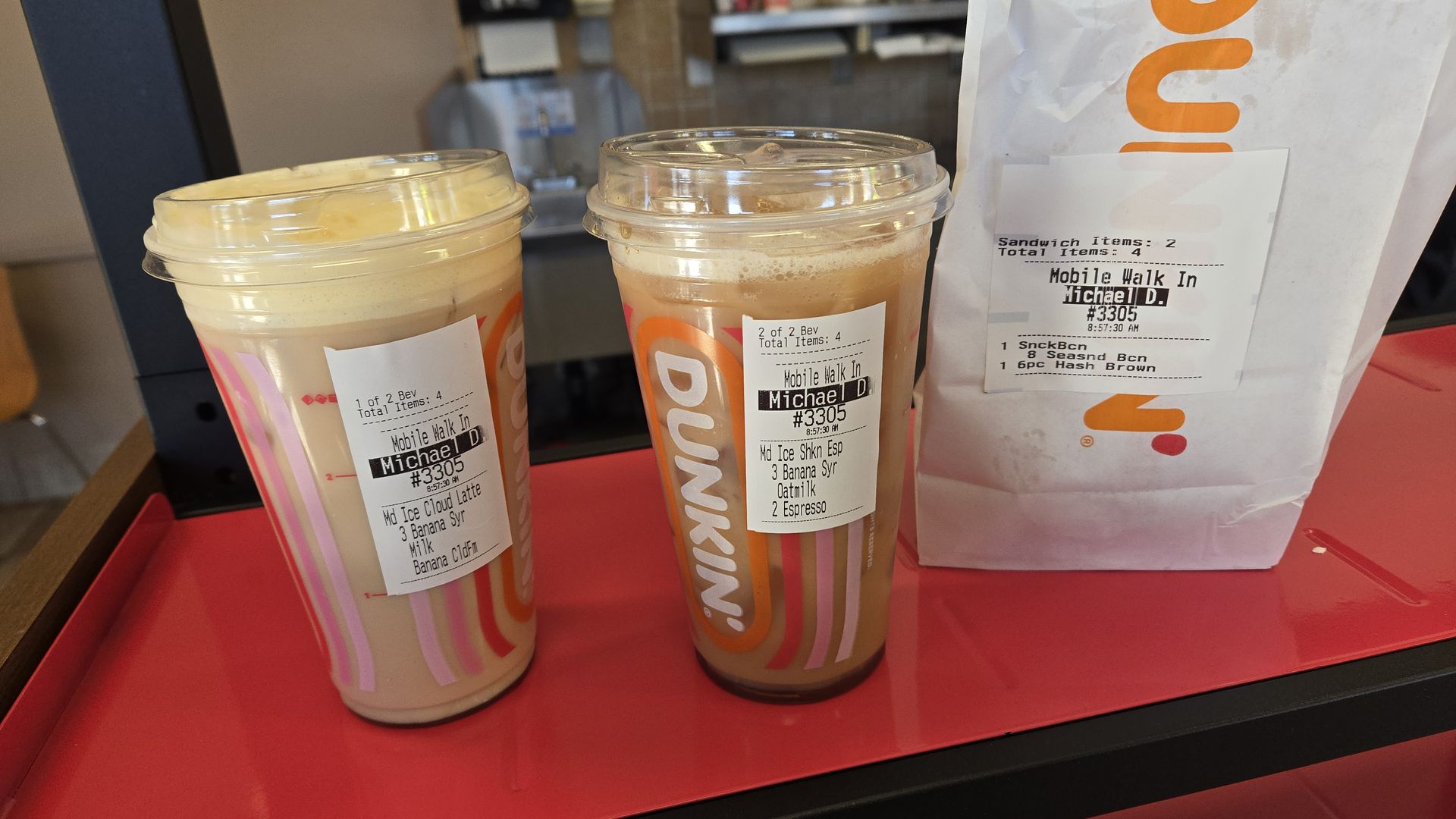 Two Dunkin' iced coffees on a red tray with attached order labels reading Mobile Walk In and Michael D; a white Dunkin bag with orange logo in the background.