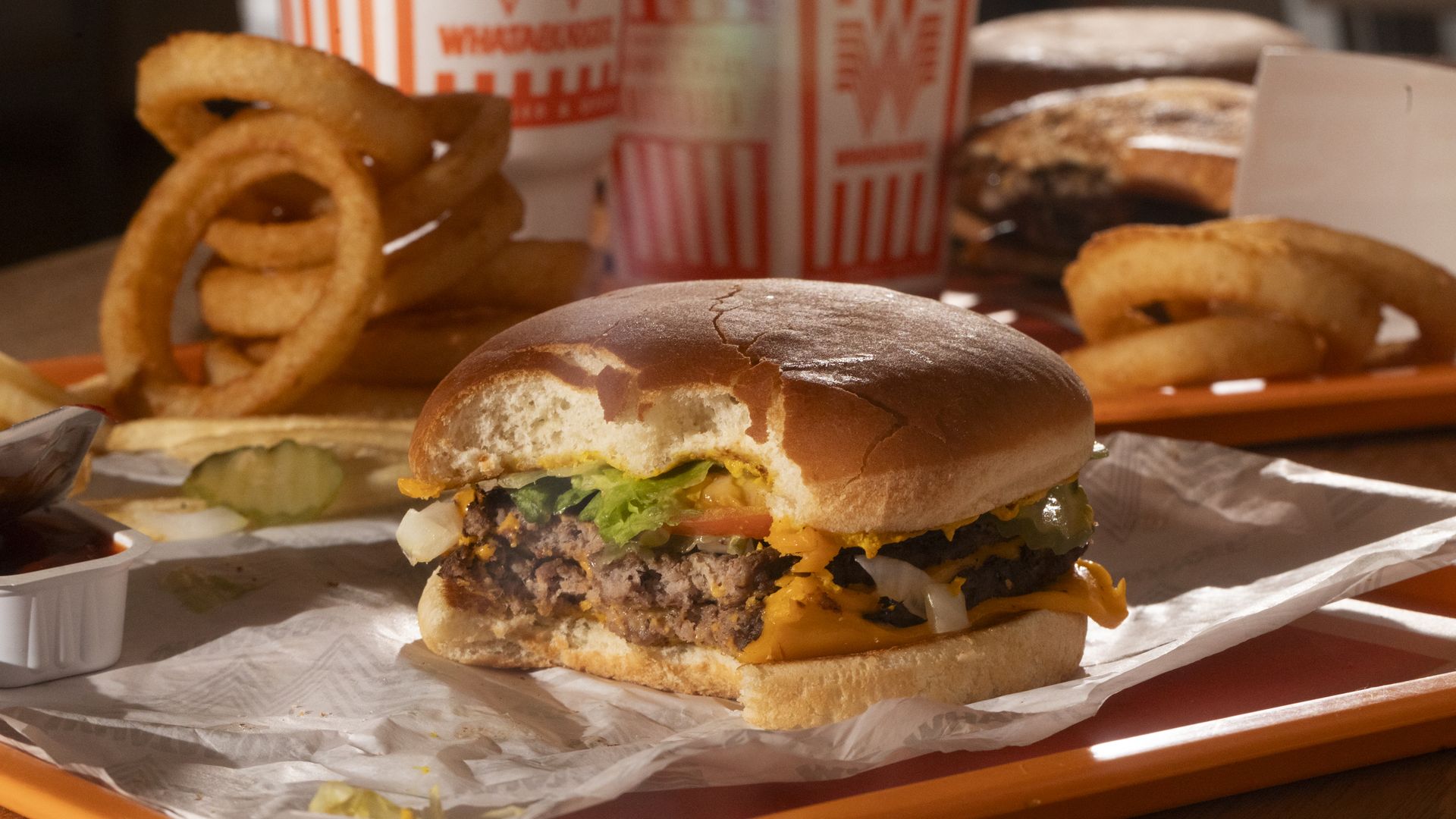 A Whataburger double meat cheeseburger is seen on a tray in front of onion rings and Whataburger-branded drink cups.