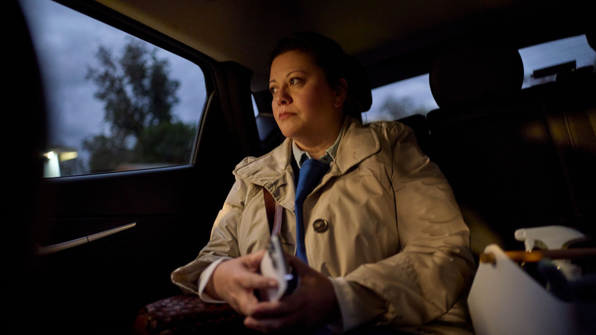 A person in a trench coat and blue tie sits in the backseat of a car, looking out the window with a neutral expression.