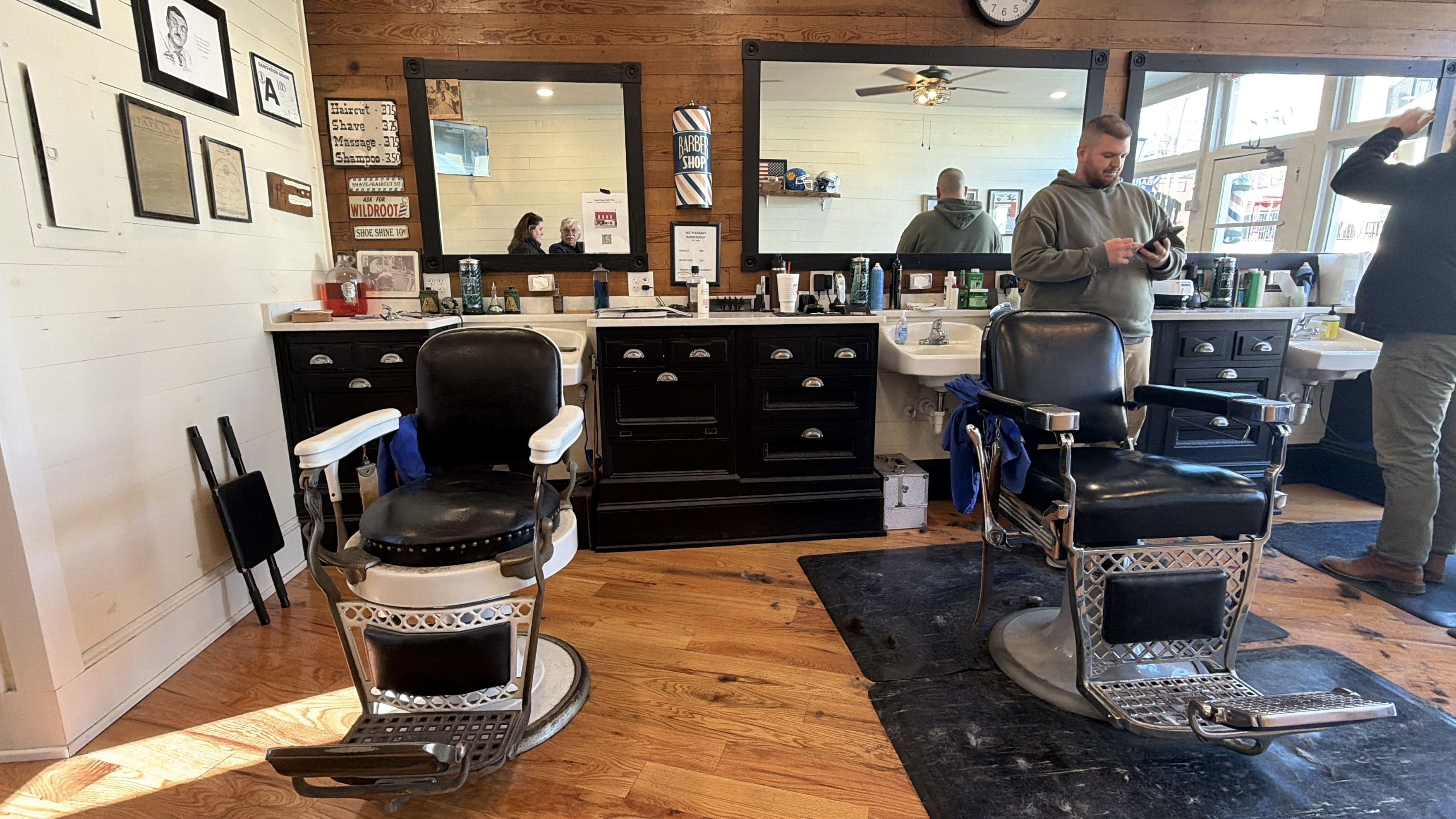 Interior of a barbershop with two vintage black barber chairs, wooden floors, large mirrors, dark wooden cabinets, and two men, one using a phone and another adjusting his hair.