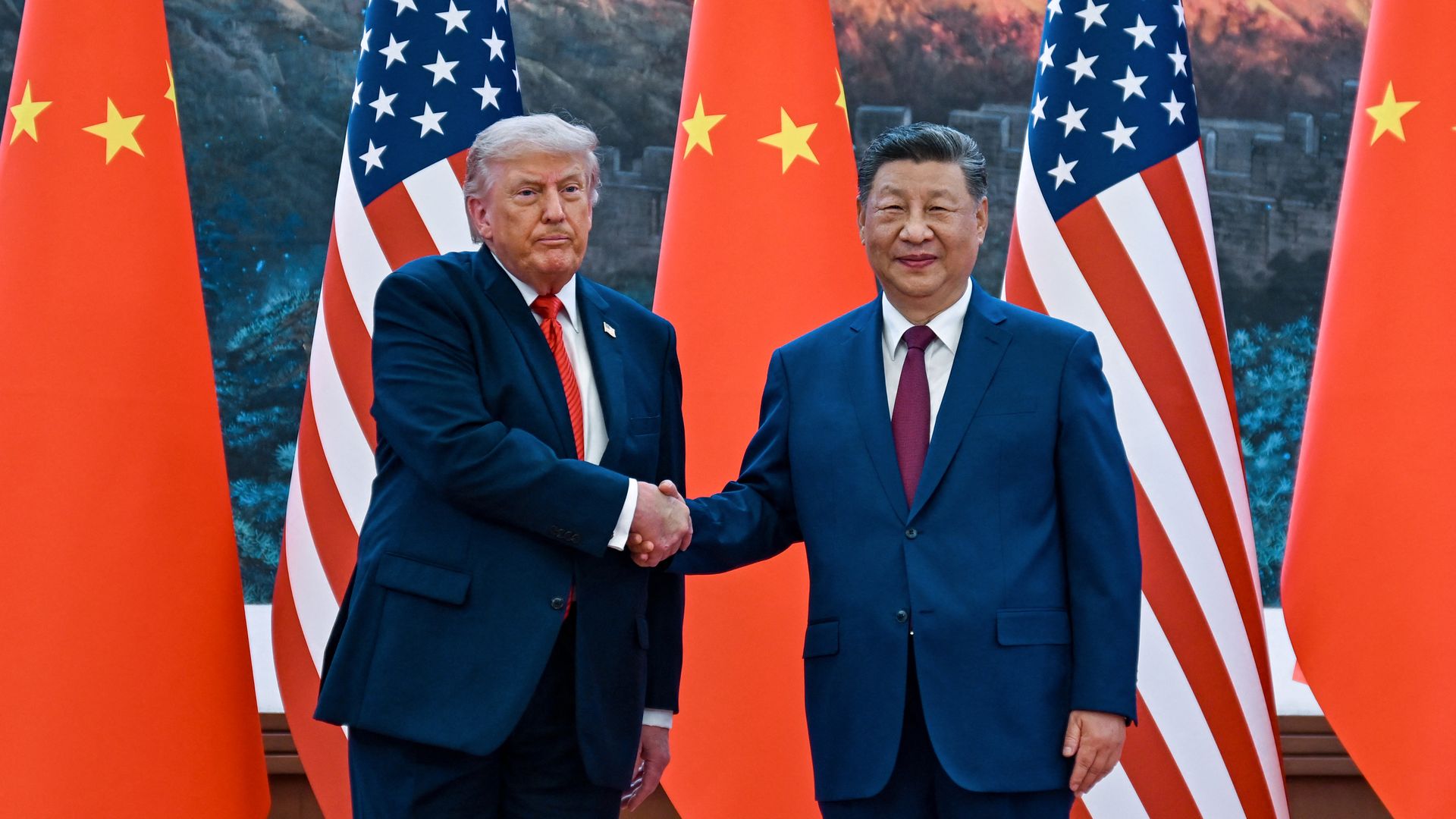 Two leaders, Donald Trump and Xi Jinping, shake hands in front of alternating US and Chinese flags with a mountain mural backdrop.