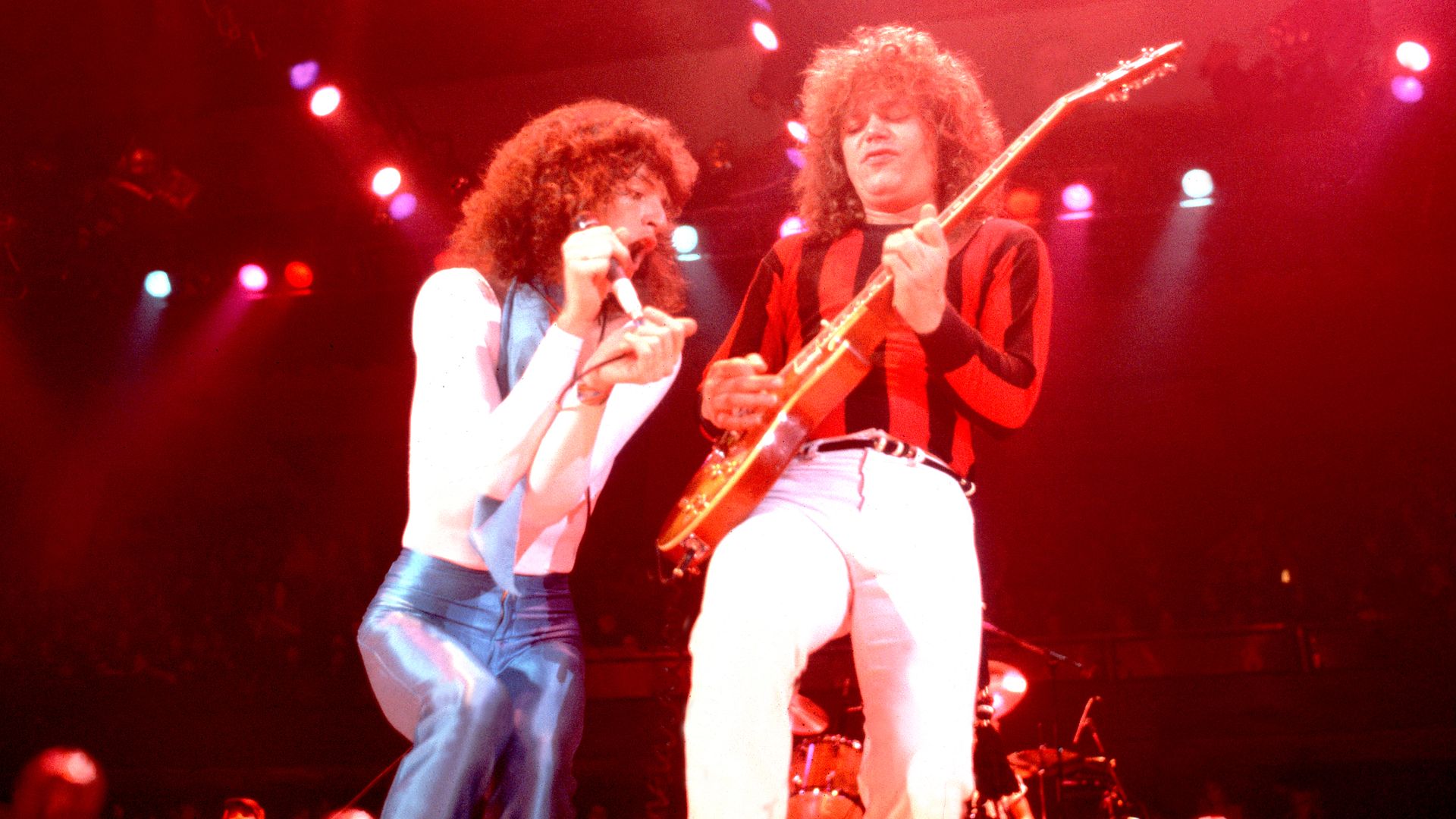 1970s white singer and guitarist, both with puffy hair, perform next to each other on stage.
