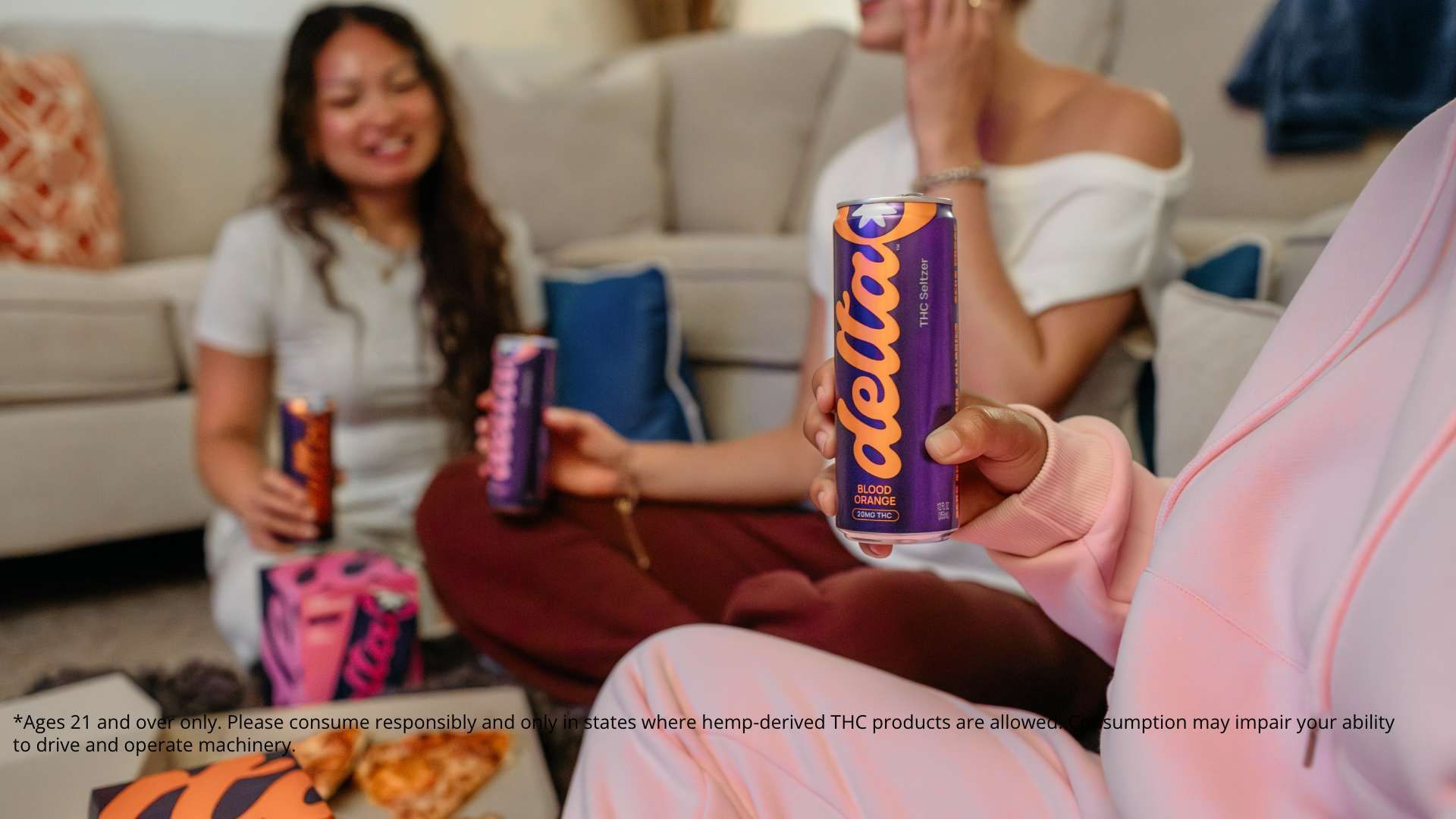 Three people sitting on a couch, casually holding purple and orange cans of Delta-8 THC seltzer labeled Blood Orange, with pizza on a table in the foreground.