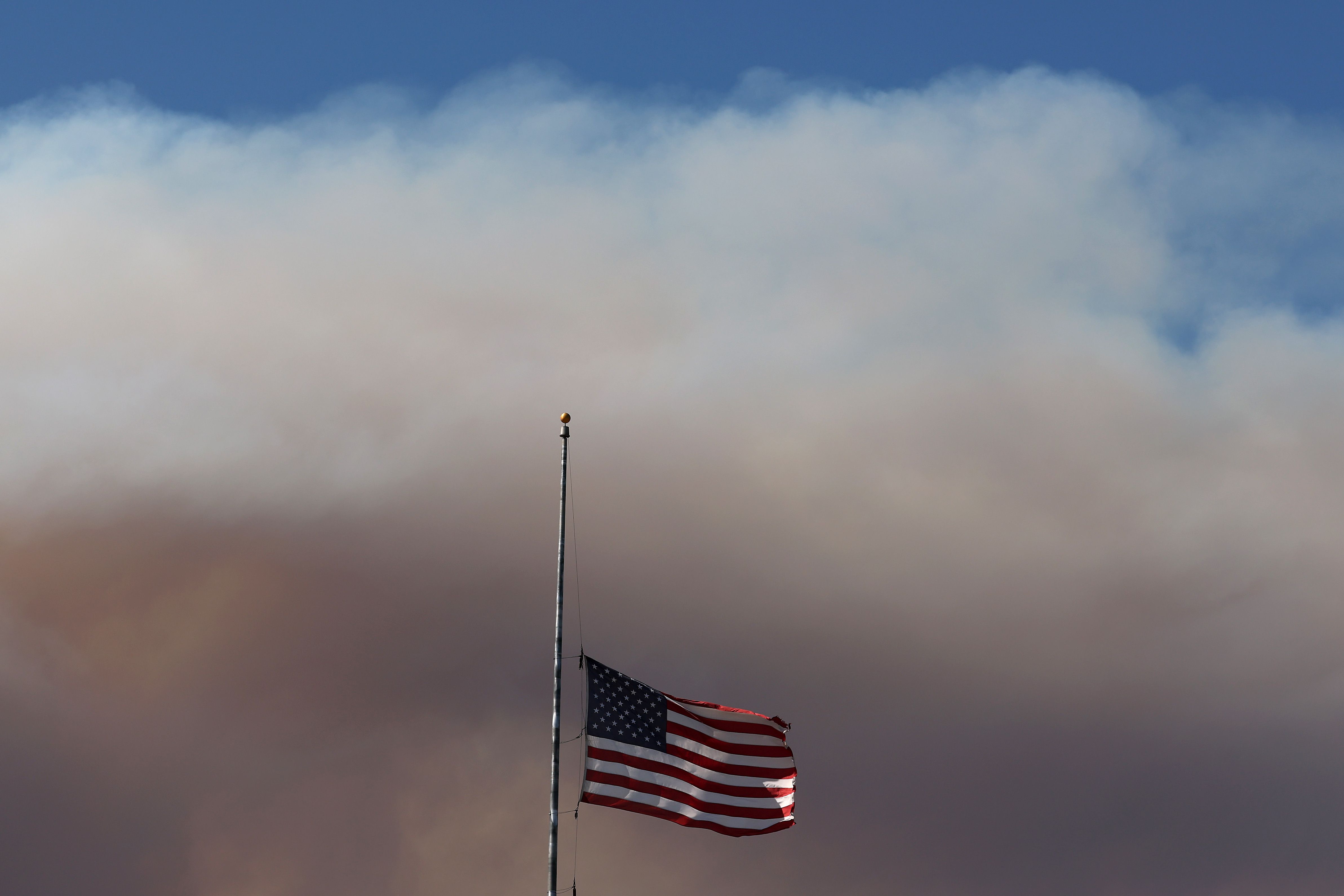 American flag flying at half-mast on a flagpole against a background of thick, dark smoke and a clear blue sky above.