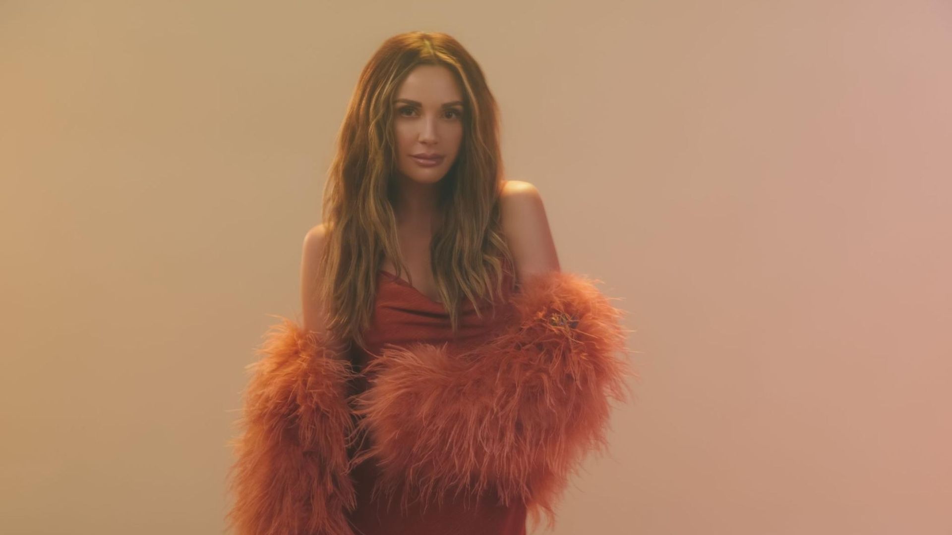 A woman wearing an orange dress stands in front of a plain background. 