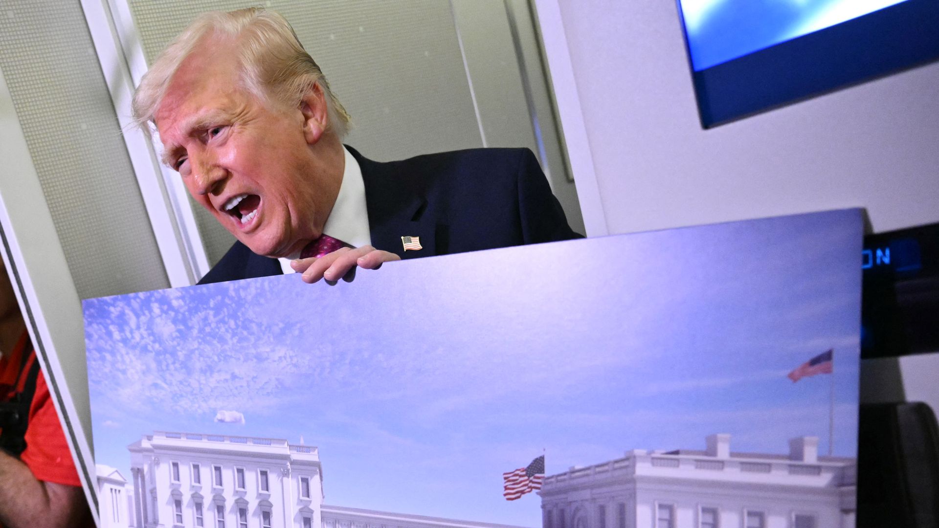 A man in a dark suit with pink tie leans over a silver barrier, mouth open as if shouting. White hair and a small American flag lapel pin are visible.
