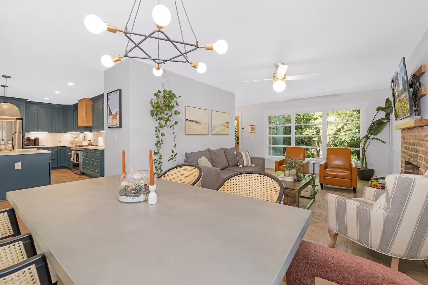 Open living space with gray sofa, two brown leather chairs, striped armchair, large window, green plants, blue kitchen cabinets, and modern chandelier lighting.