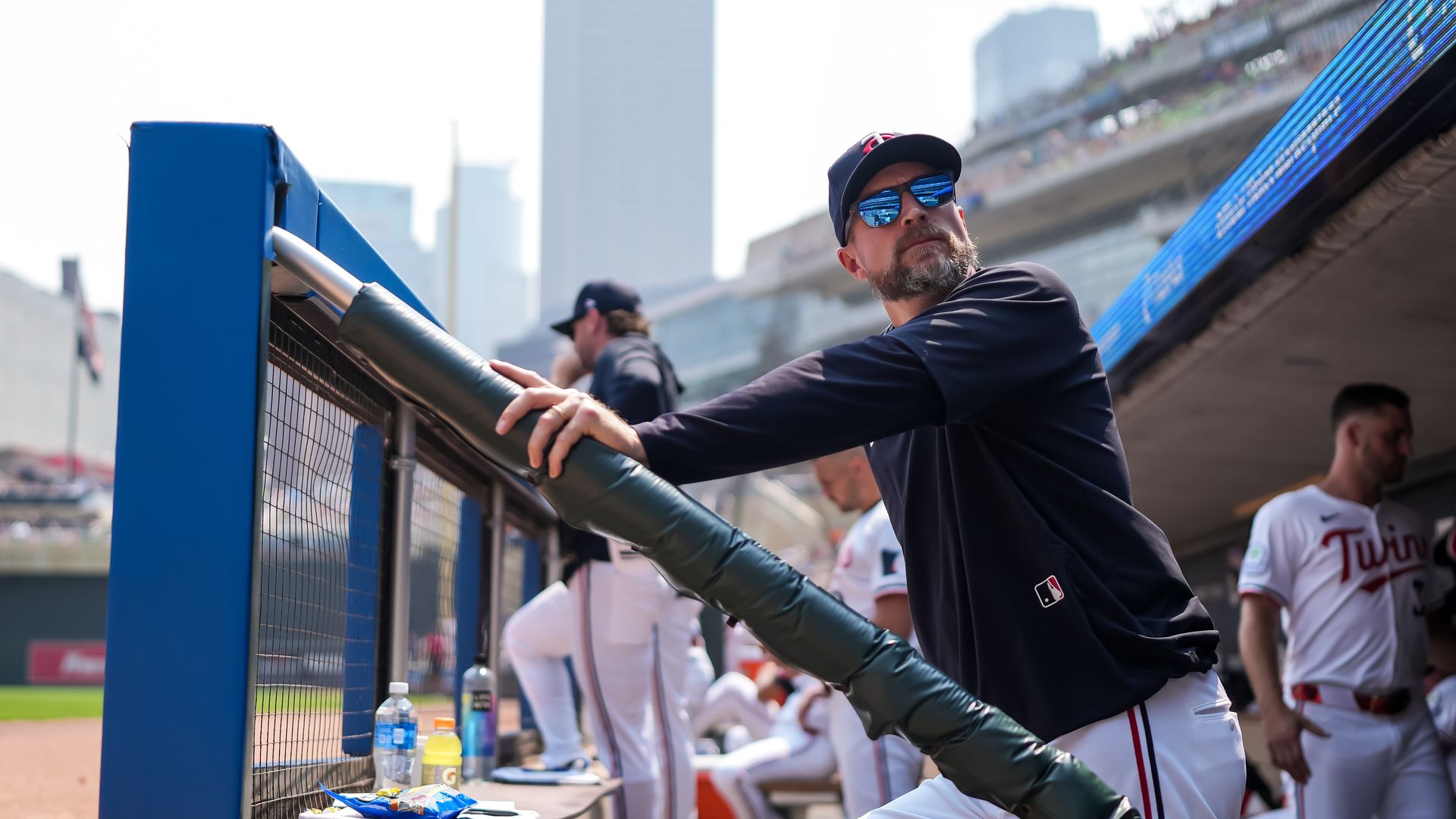 Rocco Baldelli stands in the dugout with his hand on a railing and Twins players behind him. He is wearing a hat, sunglasses, and a dark shirt. 