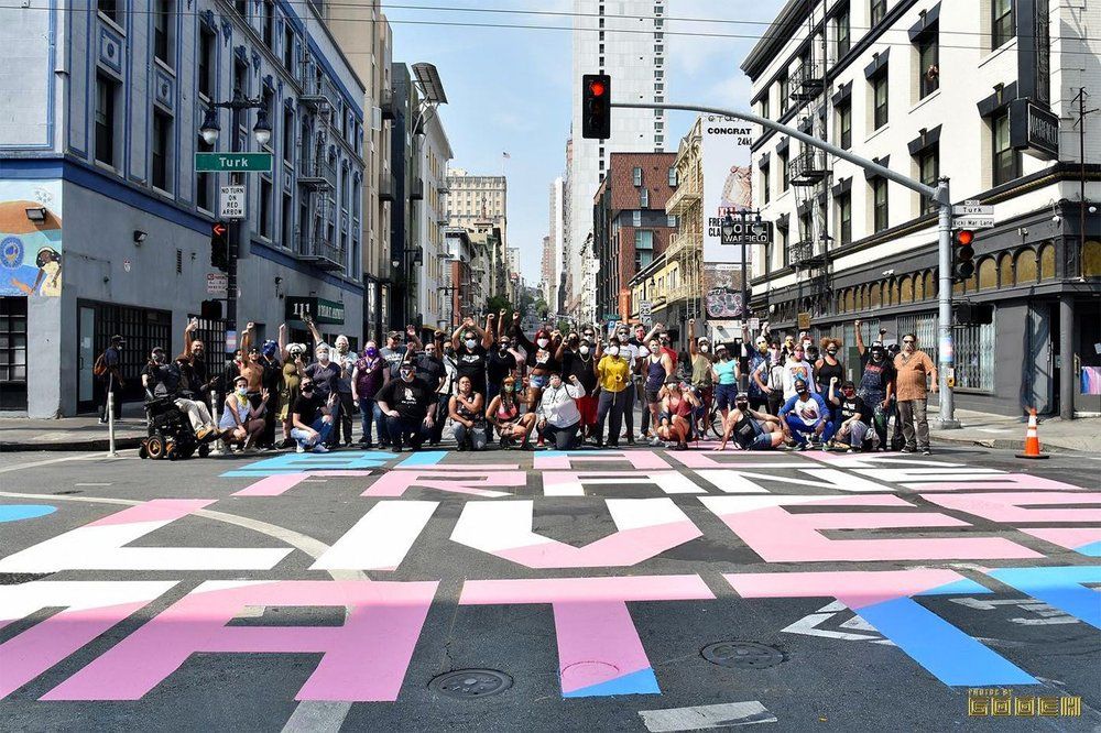 Group of diverse people, some wearing masks, pose together on a city street with large pink, blue, and white painted letters on the road reading a message, under city traffic lights.