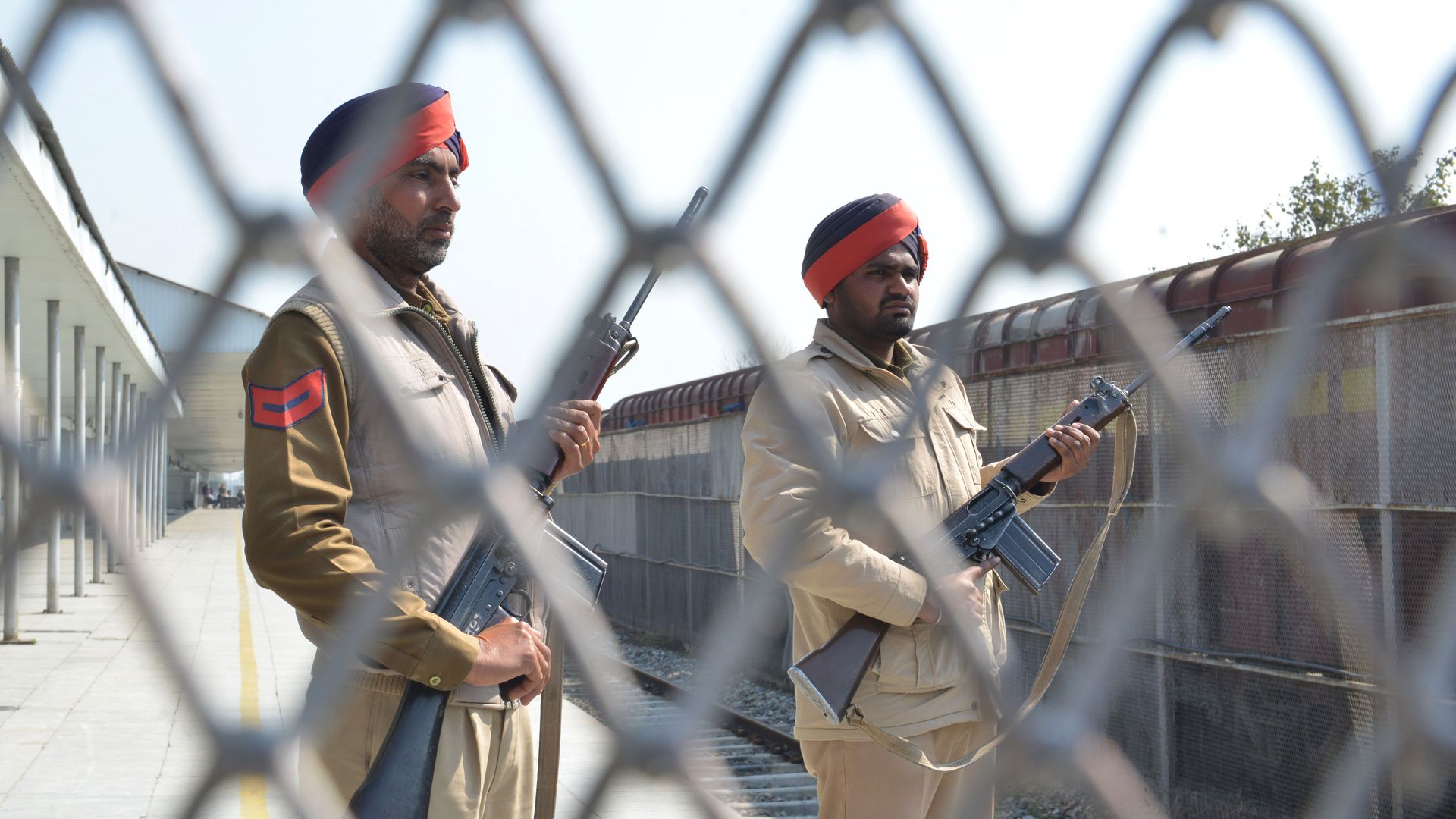 In this image, Indian Punjab Police stand guard with assault weapons behind a gated fence at a train station.