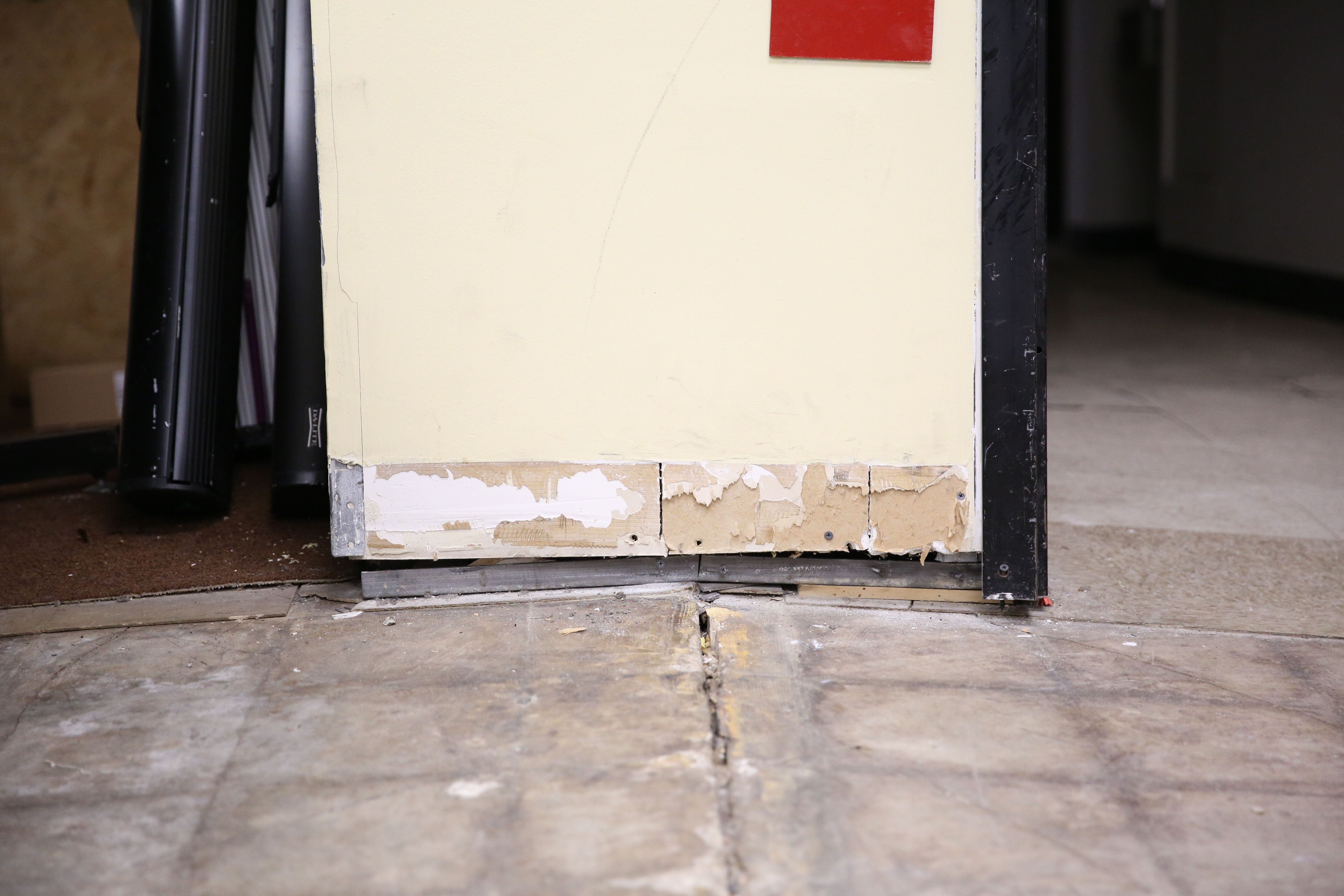 Close-up of a damaged lower wall corner with peeling paint and exposed drywall, adjacent to grey tiled and brown carpet floors, showing wear and cracks at the base.