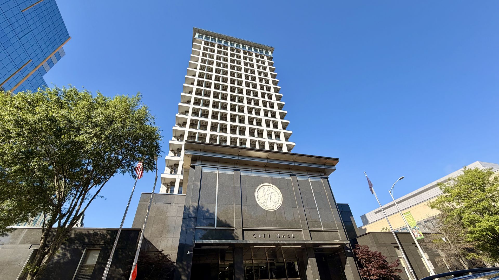 Low-angle view of a tall white high-rise with grid balconies beside City Hall, a seal on the dark facade, an American flag, and green trees under a bright blue sky.
