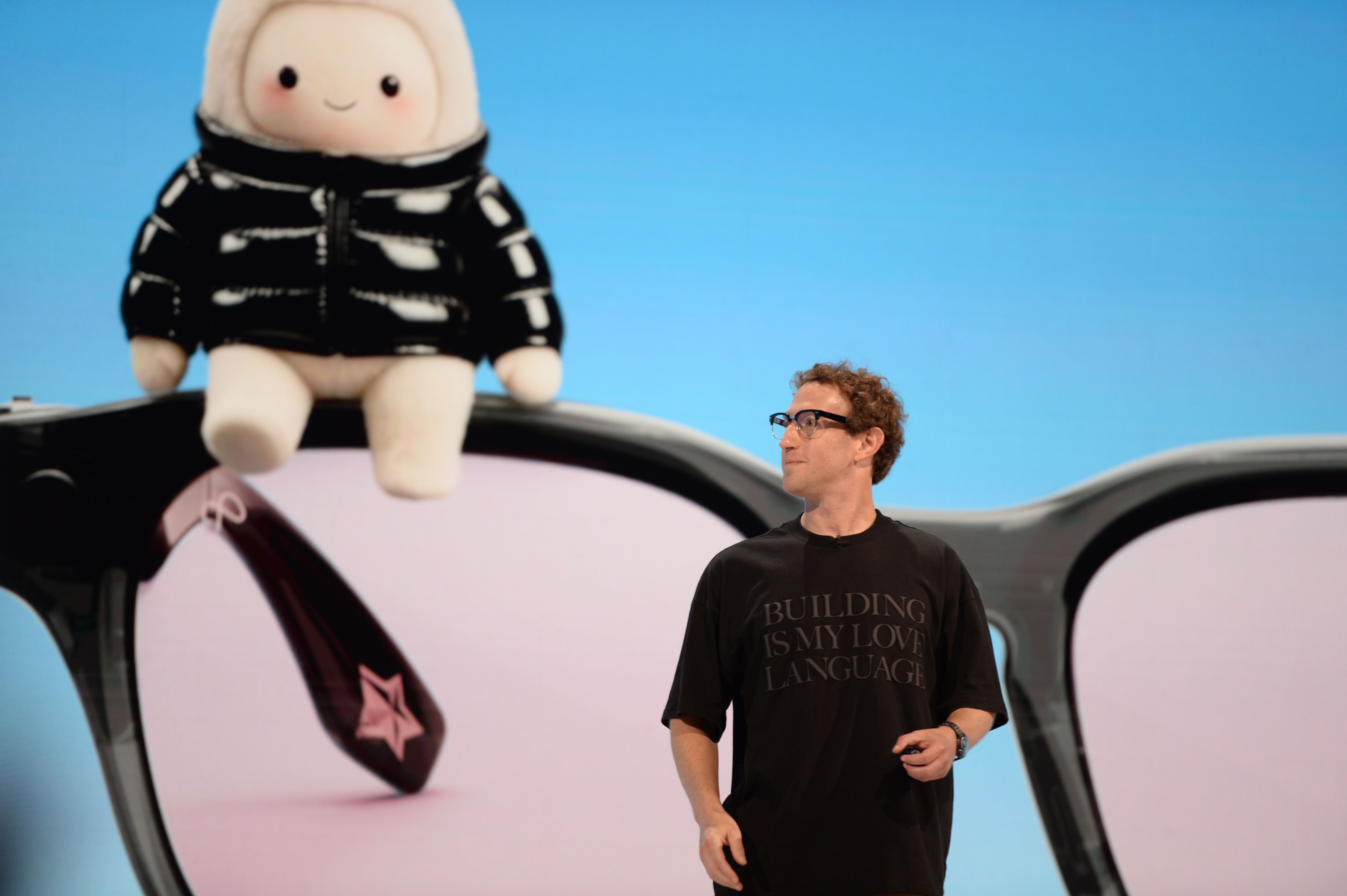 A man with curly hair and glasses stands wearing a black T-shirt (Mark Zuckerberg) stands in front of a screen displaying a mascot wearing a black jacket sitting on a pair of glasses against a light blue background.