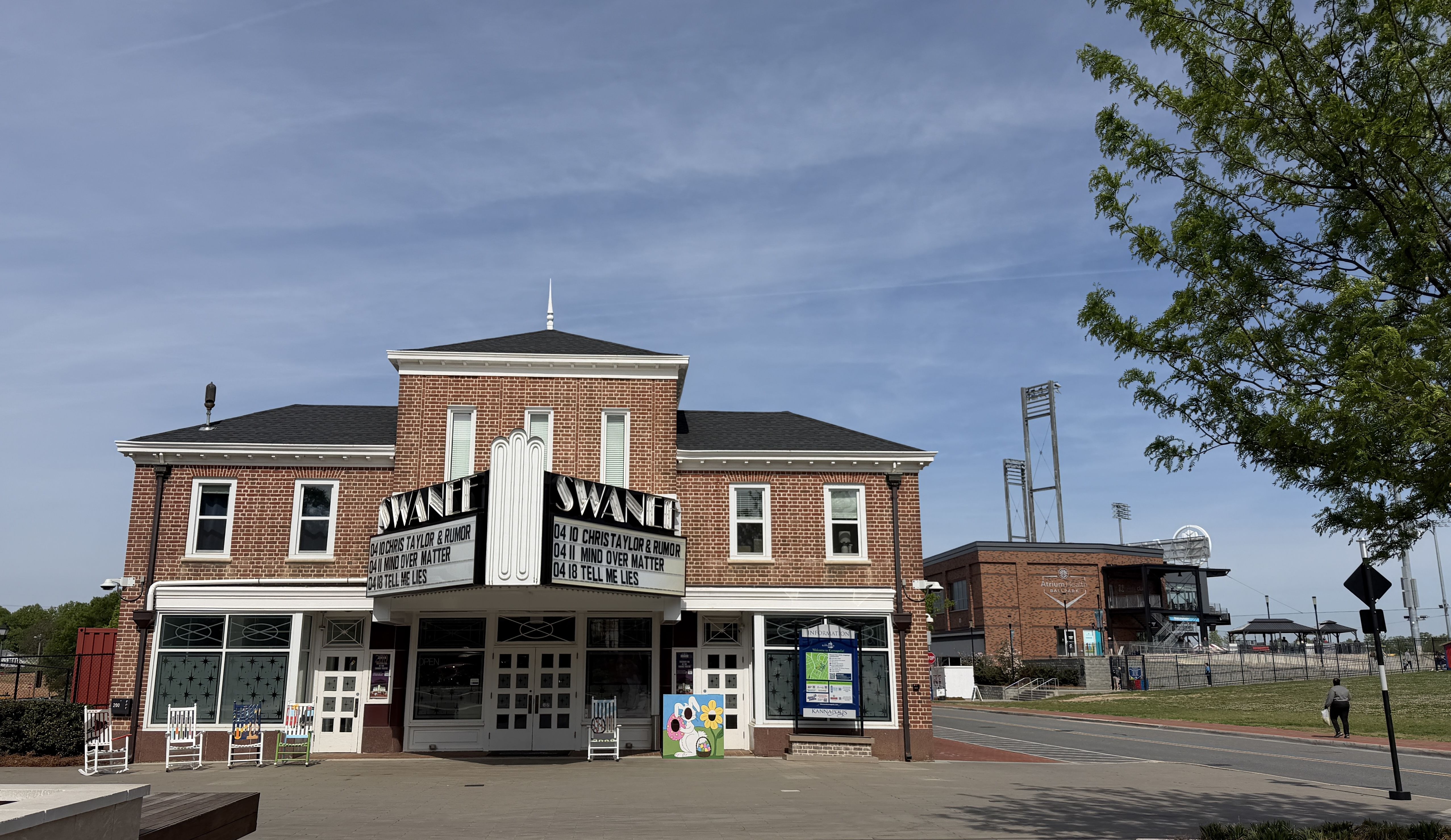 Red-brick, two-story theater with a vintage marquee reading "SWAN" above double doors; white trim and large windows. Blue sky, a tree on the right, and a street with a distant sports complex.