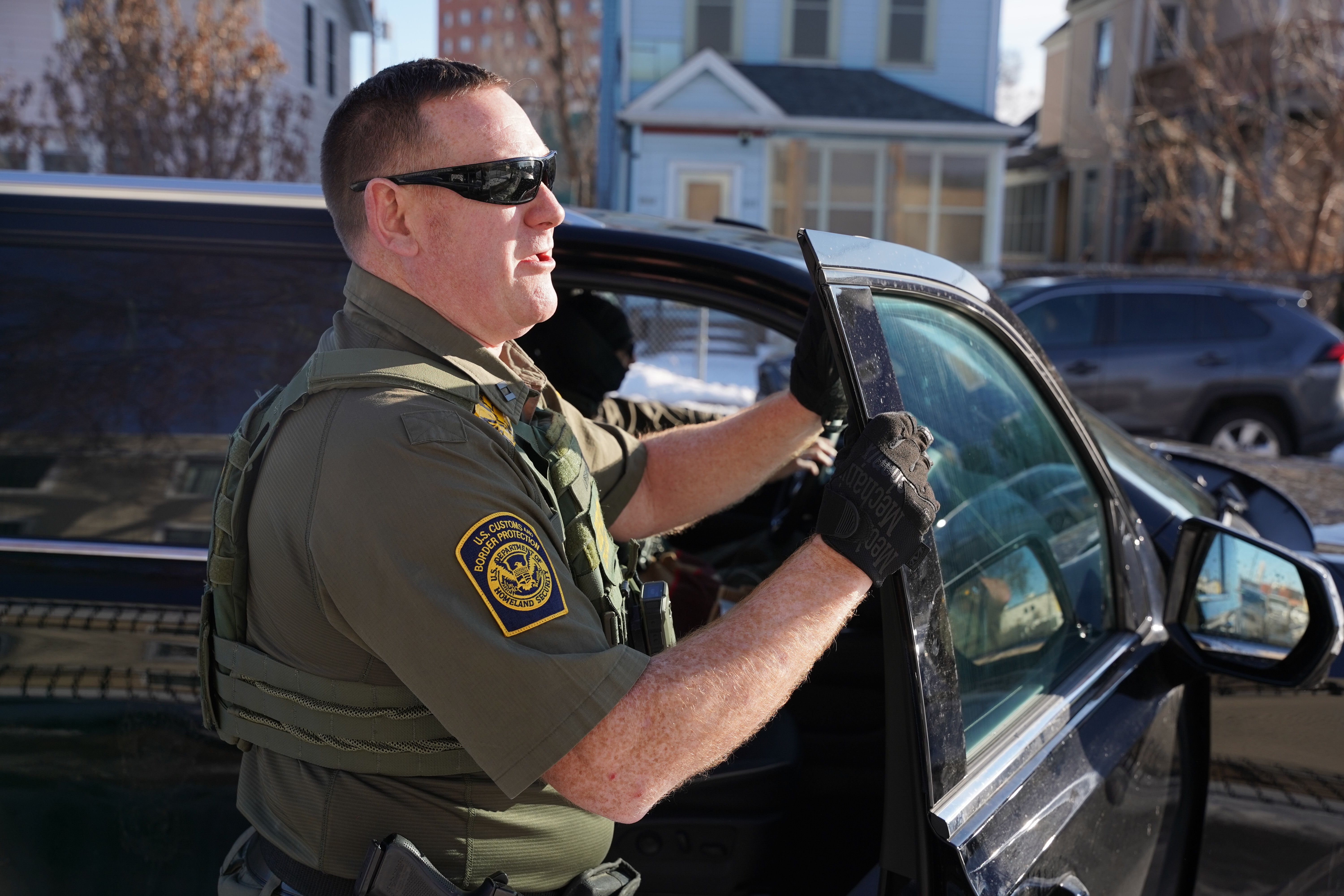 A U.S. Customs and Border Protection agent stands in a street in south Minneapolis where federal immigration enforcement activity is underway, with law enforcement vehicles and personnel visible in the background.