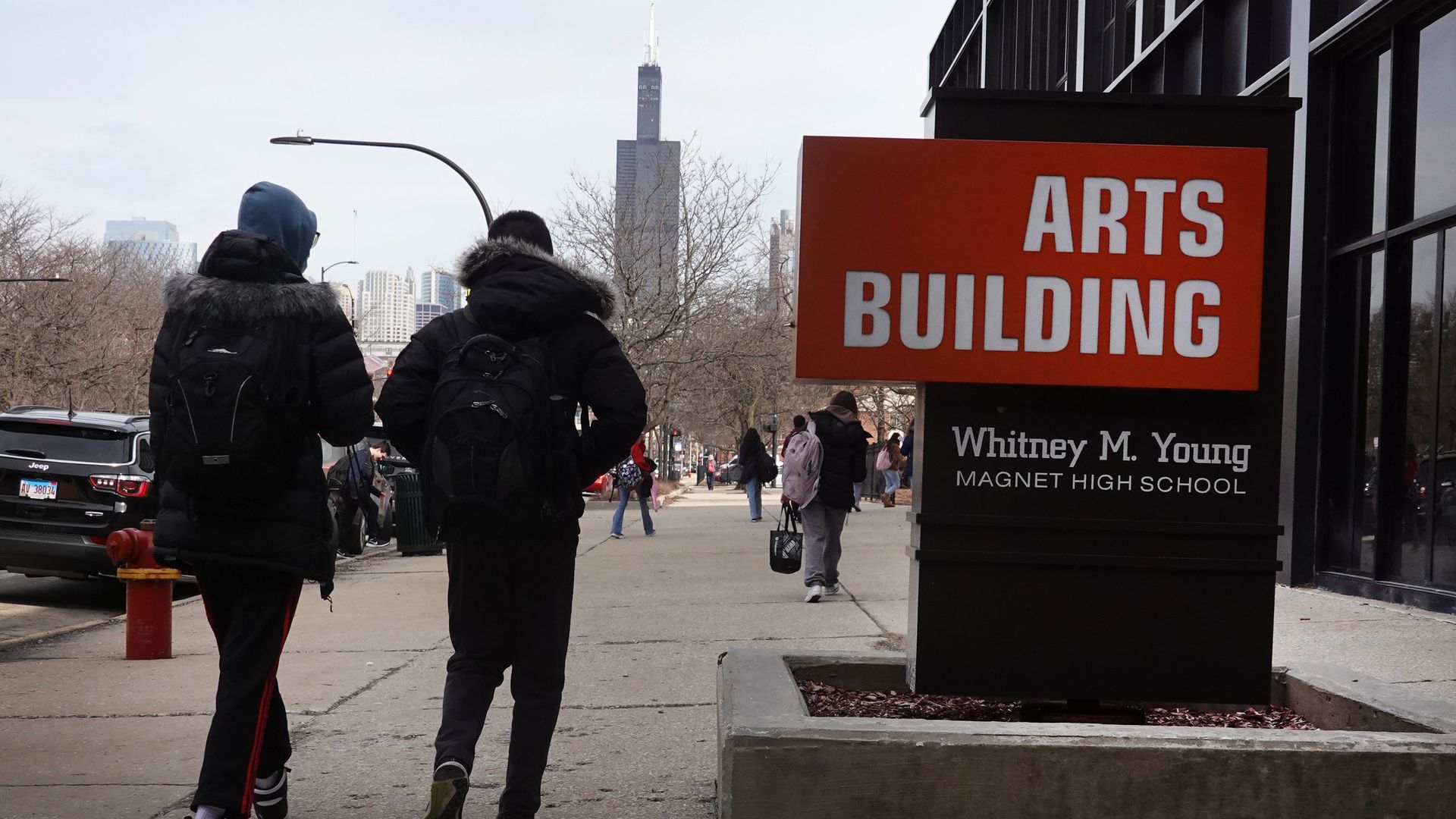Two people in black puffer jackets with fur hoods walk away on a city sidewalk. A large orange sign reading "ARTS BUILDING" sits near a glass-front building; bare trees and skyscrapers in winter.