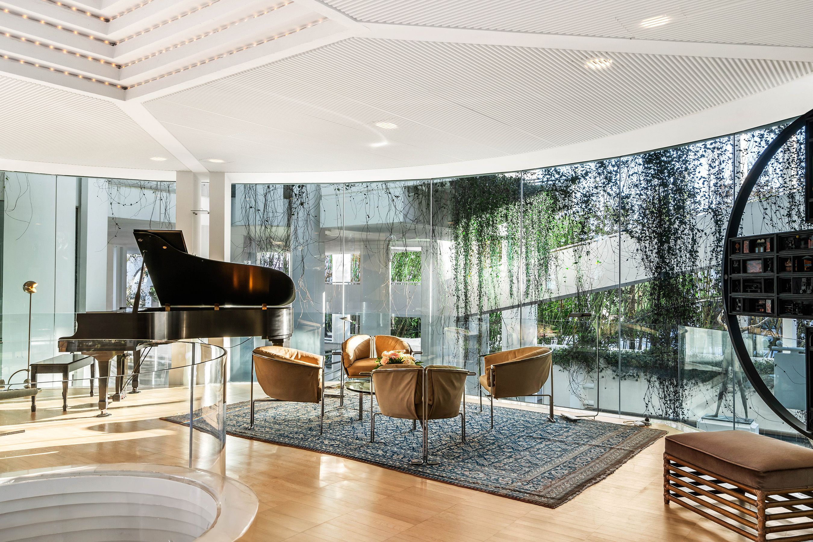 A photo of a grand piano and designer furniture in a window-filled room overlooking a ramp covered in vines