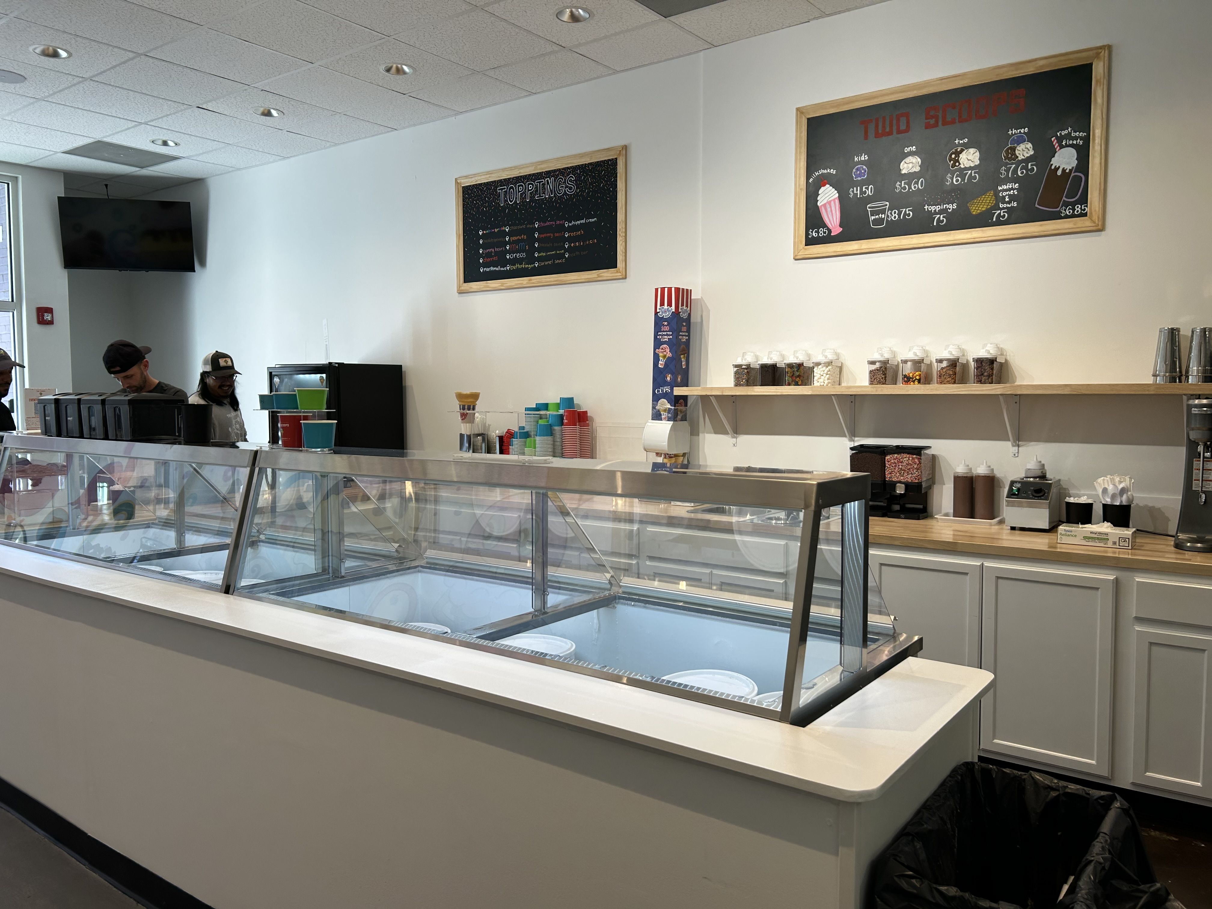 Interior of ice cream shop two signs showing toppings and prices, glass counter with no ice cream visible, shelves with toppings jars, customers ordering at counter.