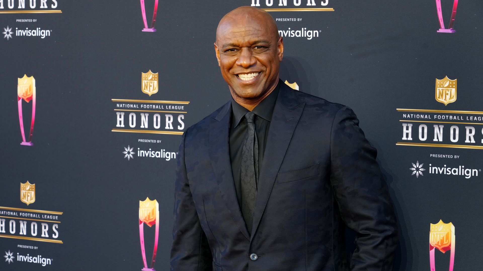 Smiling bald man in a dark suit and black tie poses on a step-and-repeat backdrop at the NFL Honors event, with Invisalign logos and pink trophy graphics behind him.