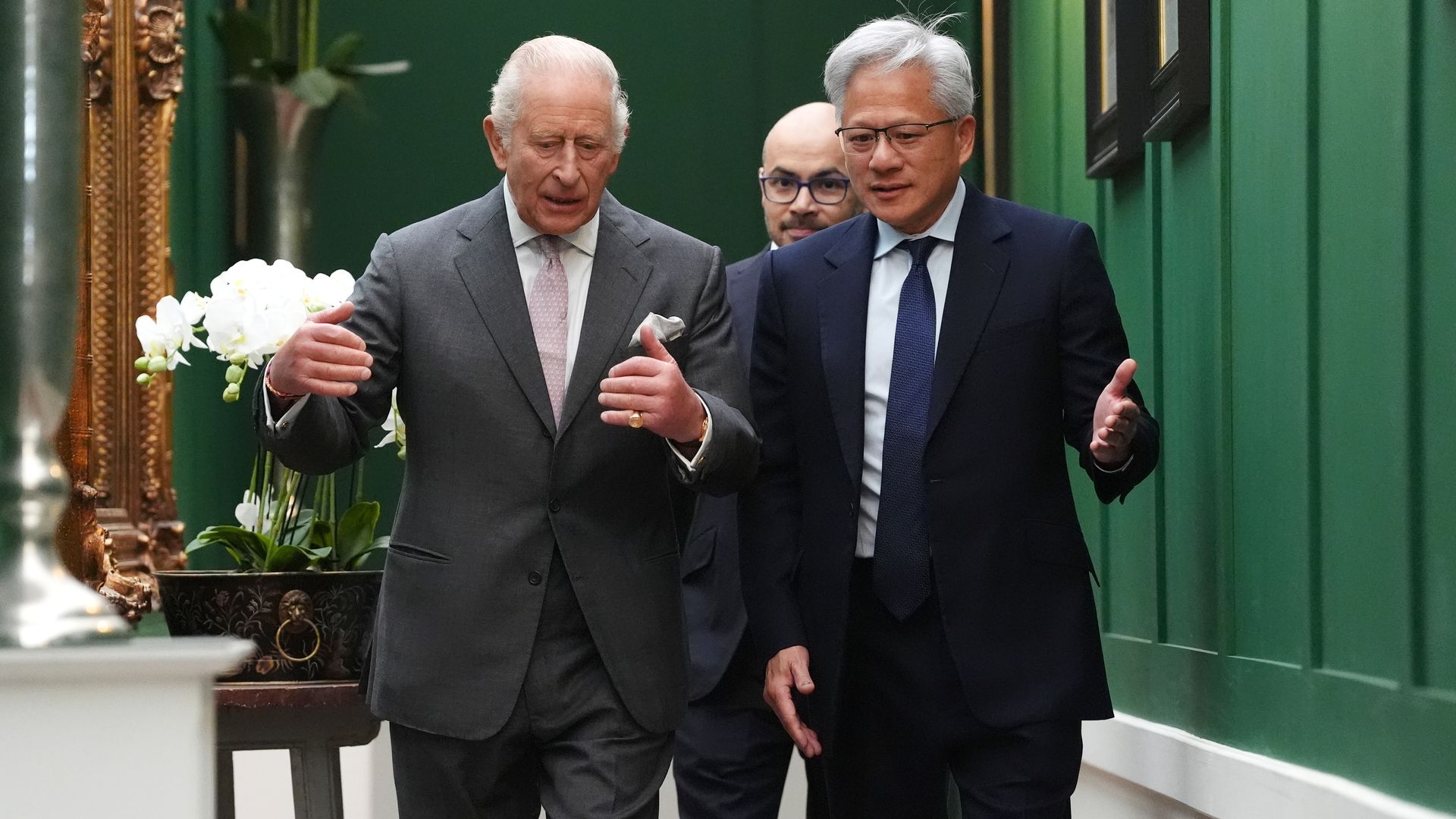 Two people in suits walk a green hallway; one in a gray suit with a pink tie, the other in a navy suit with a blue tie. A third person follows; white orchids and a gold-framed mirror visible on the left.