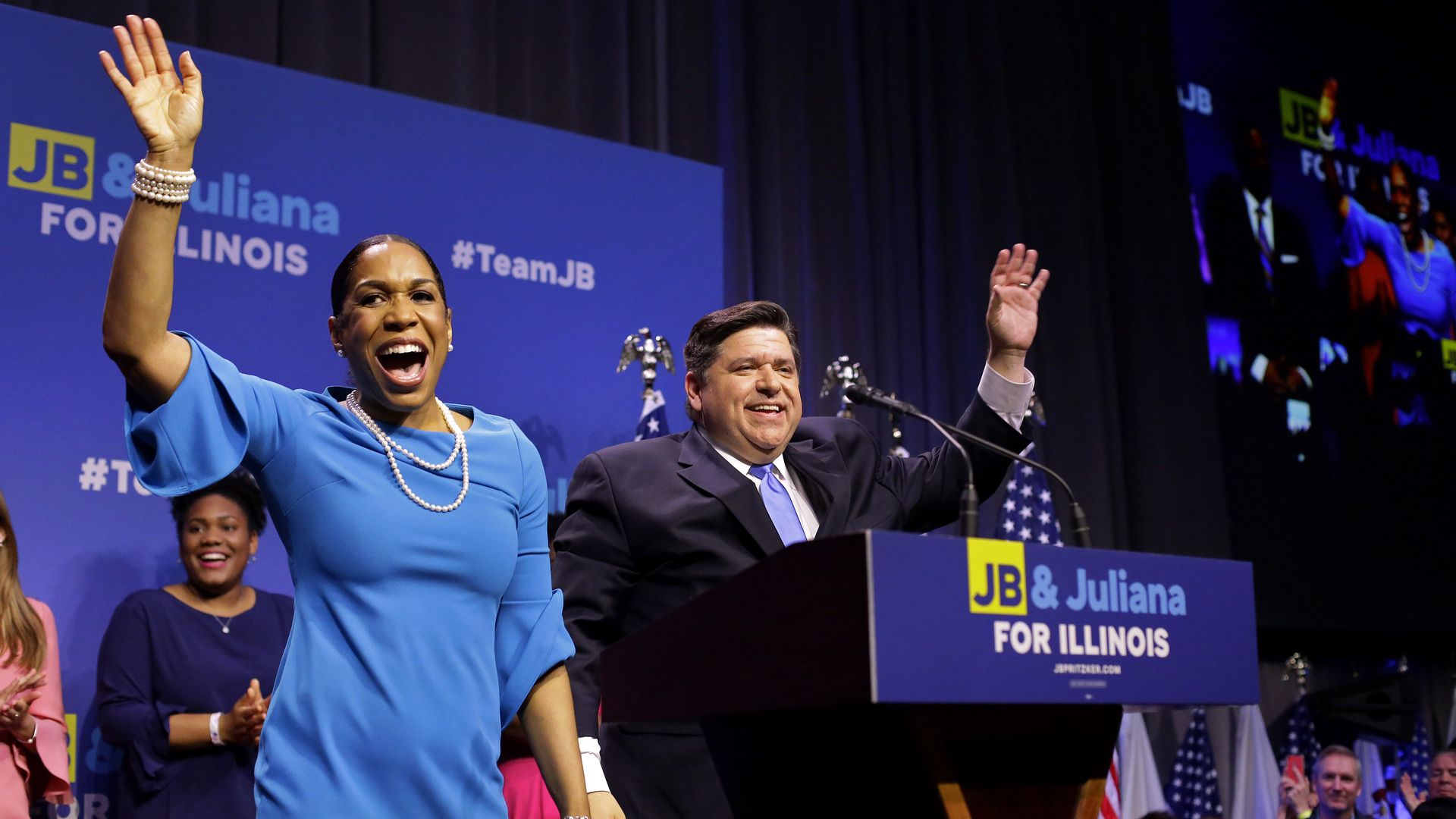 Photo of two people on a stage behind a podium waving to a crowd. 