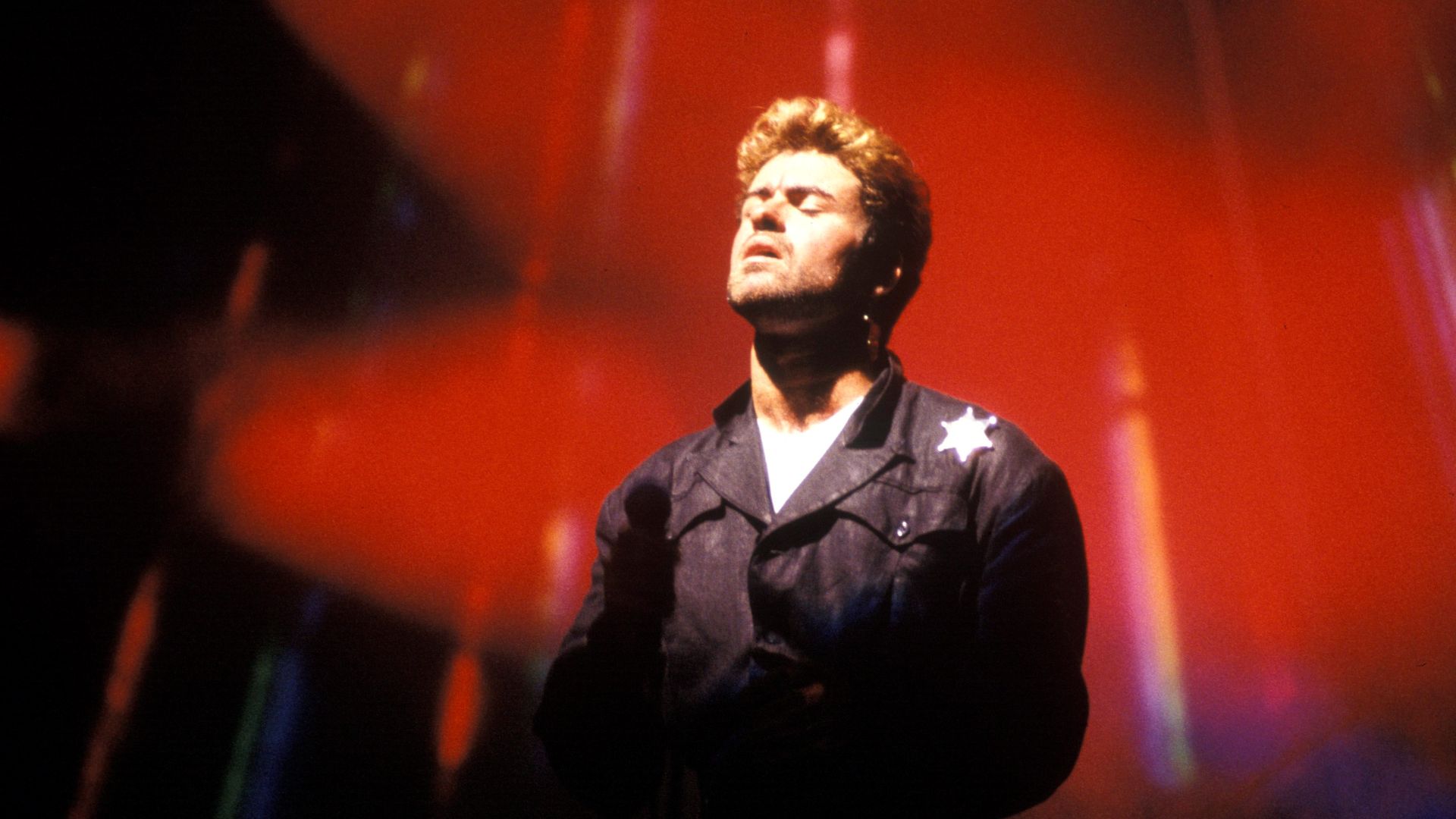 Singer George Michael stands under a re dspotlight.