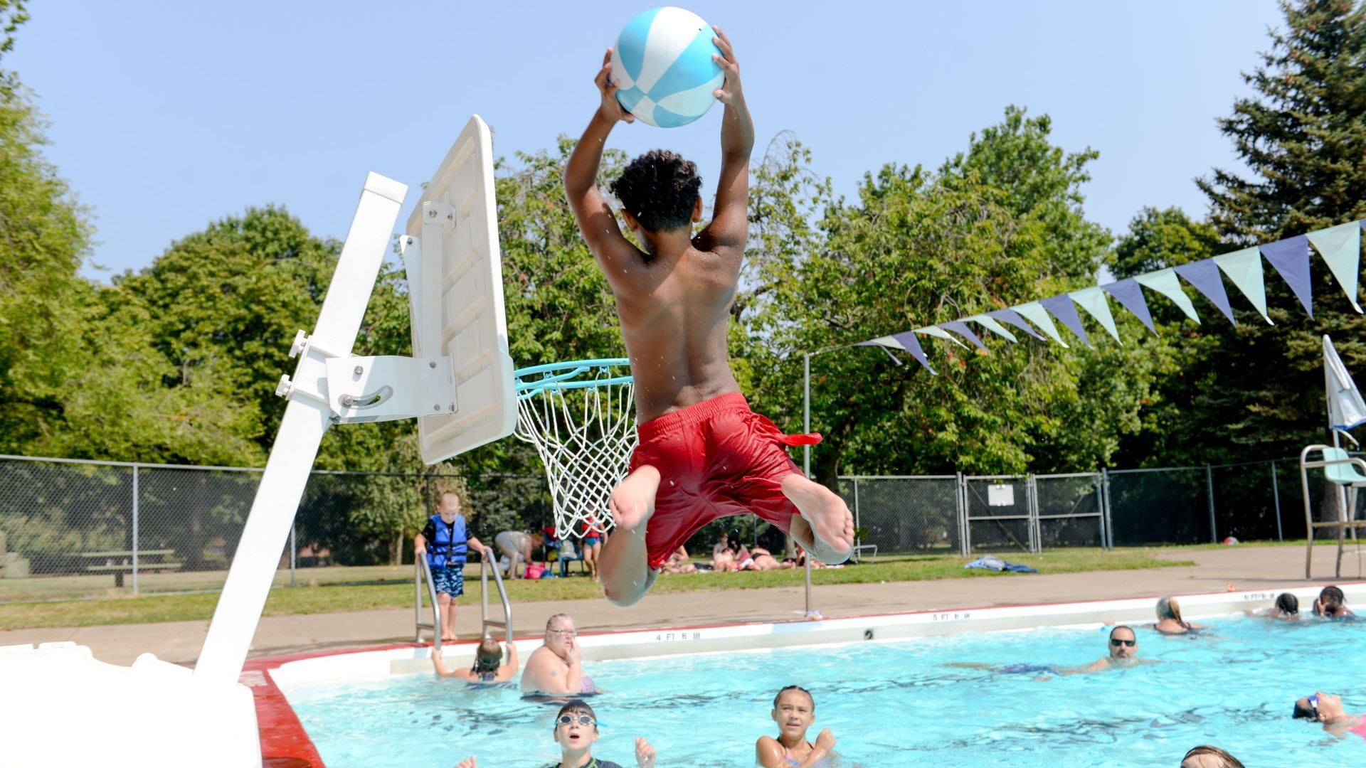 A young boy jumps high in the air to slam dunk a beach ball in a poolside basketball hoop while several other kids look on.