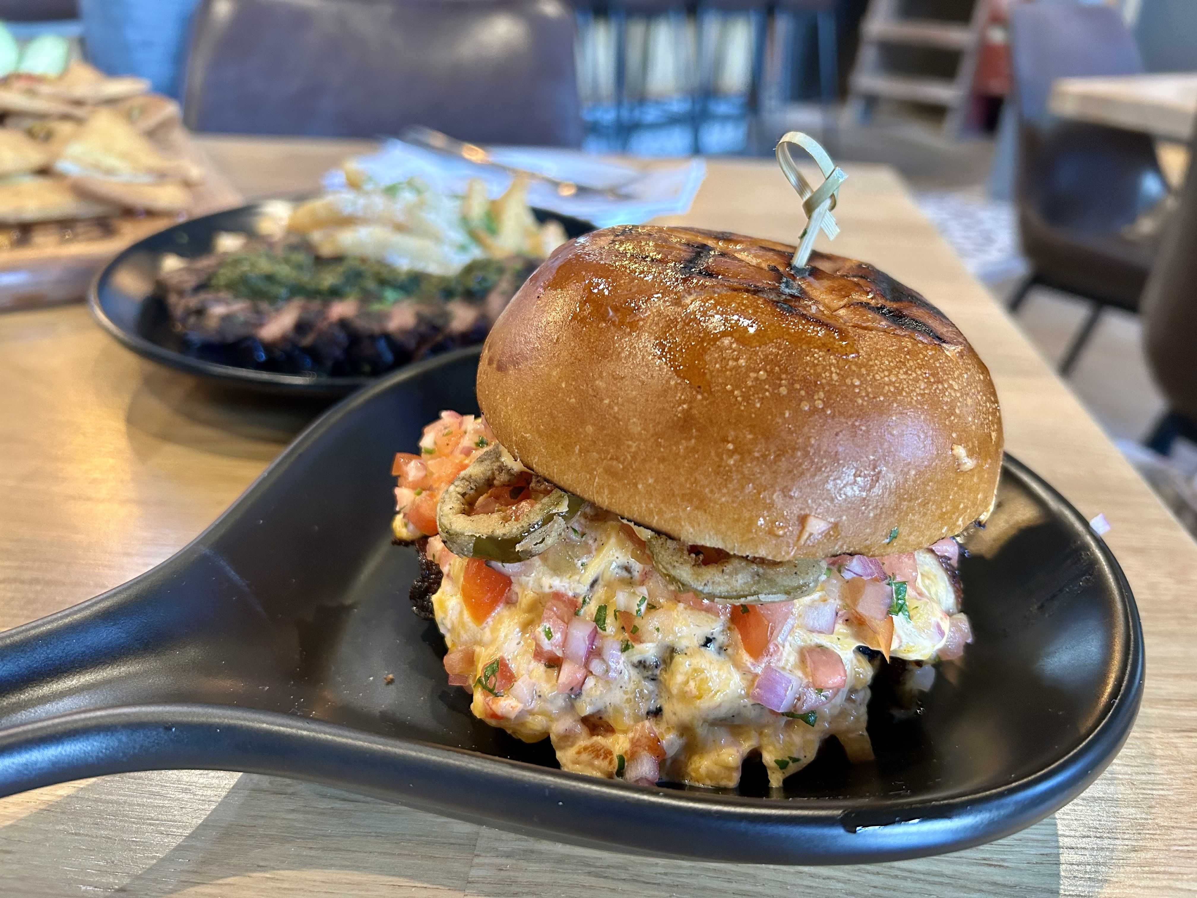 heist brewery. Close-up of a glossy bun burger with a wooden skewer through the top, stacked with a tomato-onion-herb filling on a black plate; blurred plates and tables in the background.