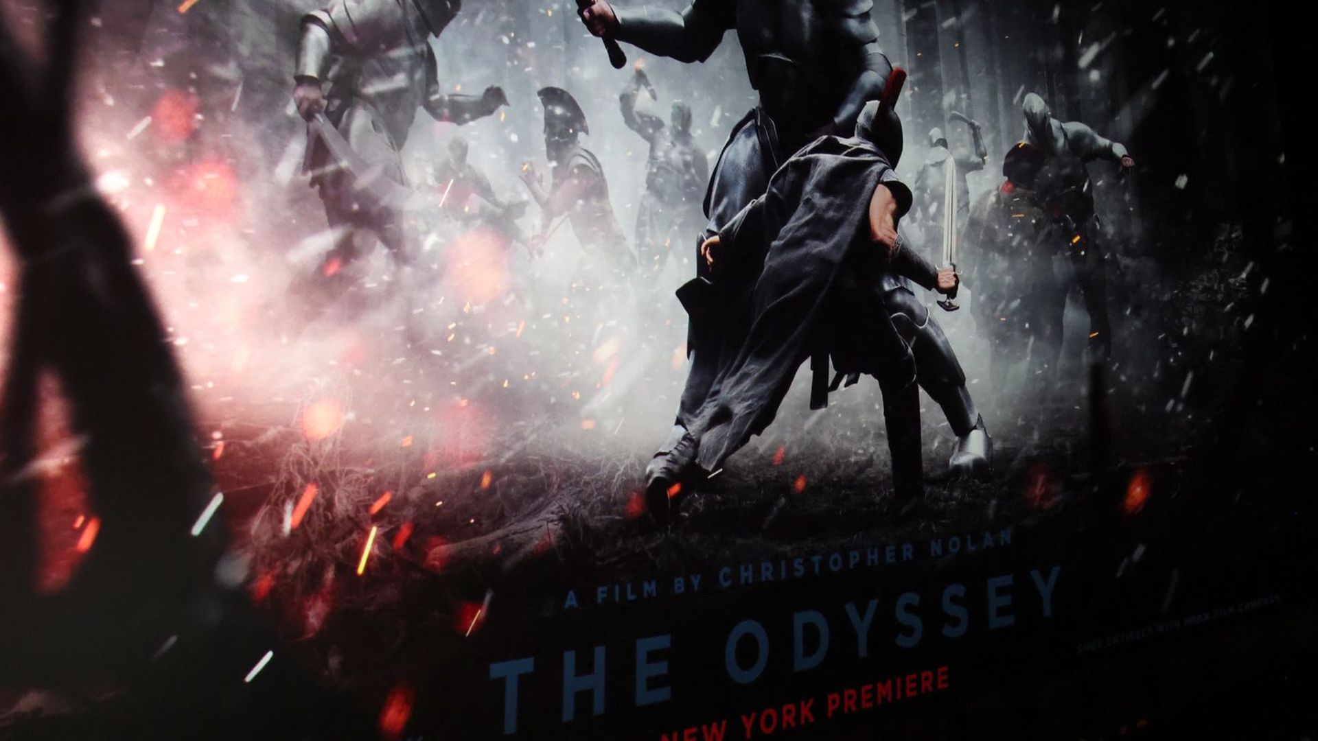 Dark movie poster depicting a dramatic sword fight: a cloaked man with a raised sword confronts armored warriors amid smoke and sparks; red lettering and the title "The Odyssey" at the bottom.