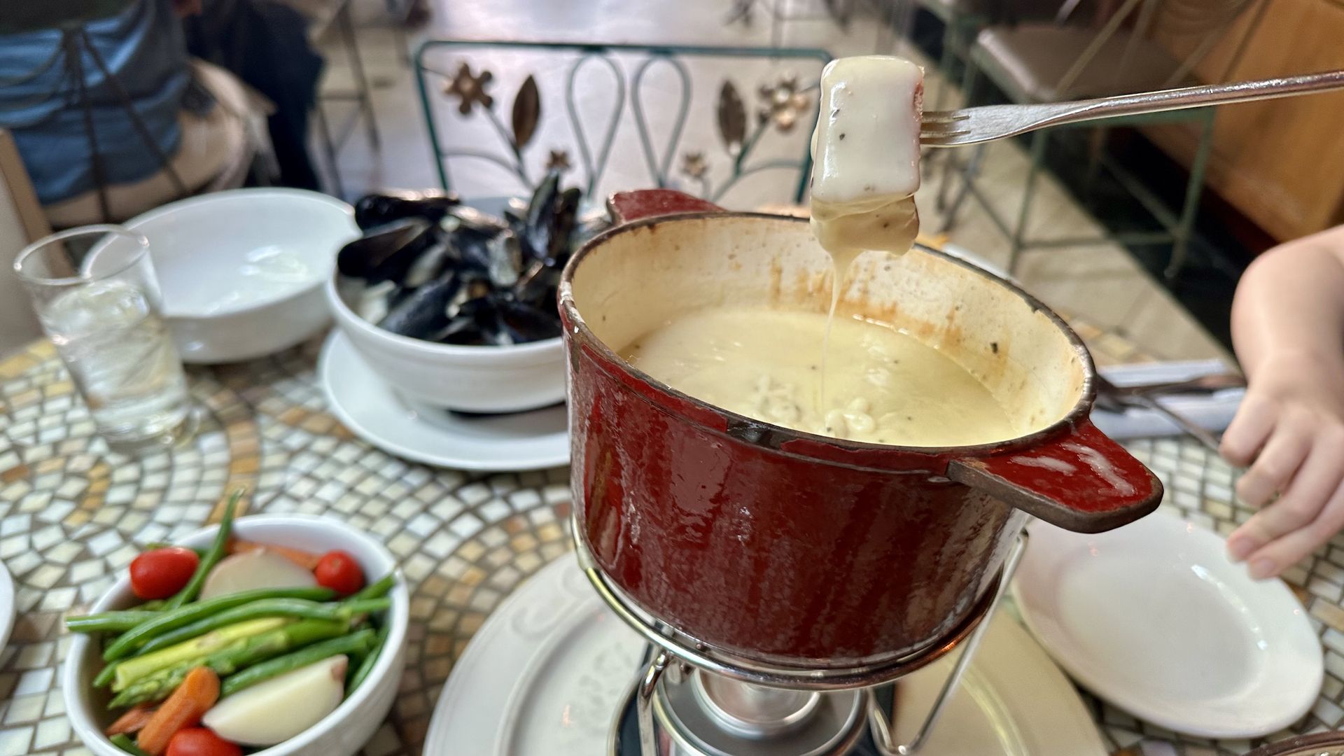 Red fondue pot on a stand with melted cheese. A fork holds a bread cube dipped in cheese. In the background, a vegetable platter and a glass of water sit on a mosaic table.