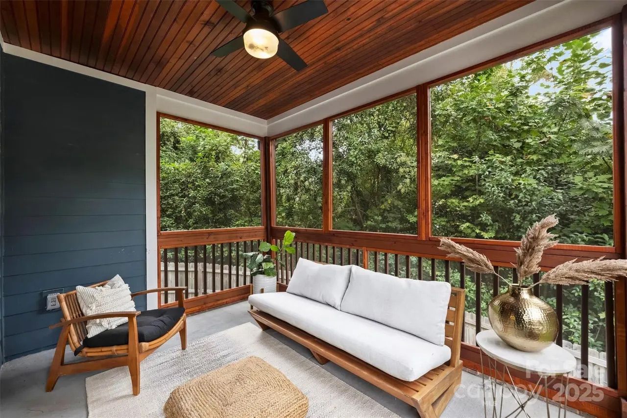 Cozy screened porch with wooden ceiling and fan, featuring a wooden chair with black cushion, a wooden sofa with white cushions, a woven ottoman, and a gold vase on a small table, surrounded by greenery.