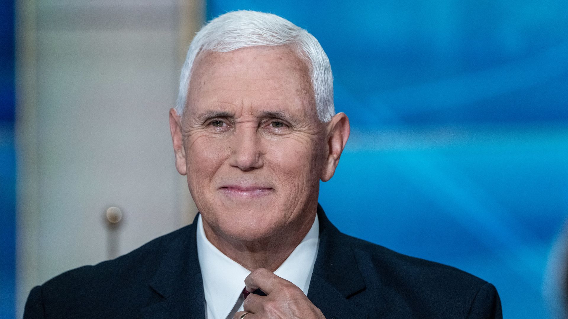 A white-haired former Vice President Mike Pence, wearing a black jacket and white shirt, smiles as he adjusts his tie, the color of which is obscured by his hand, in front of a blue background.